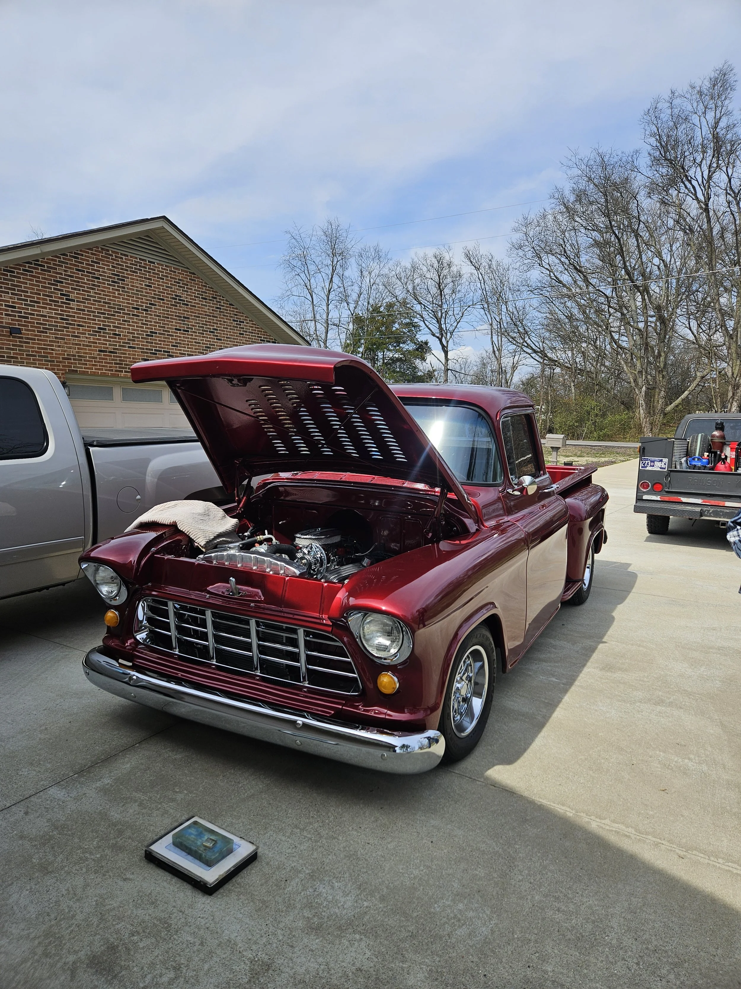 A maroon vintage car with its hood open, parked in a driveway next to a silver truck, with trees and a brick house in the background.
