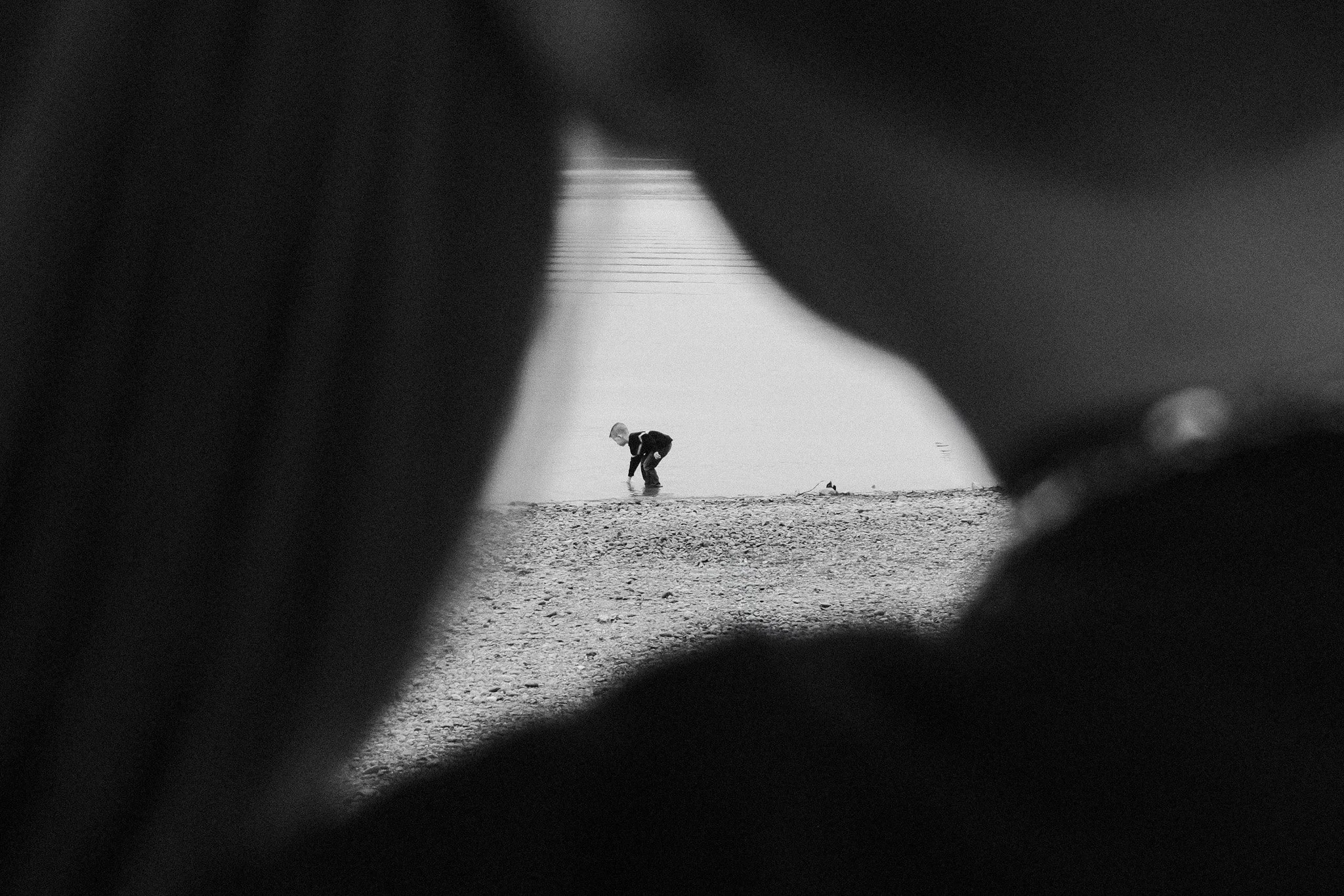 A person in a black jacket is bending down on a rocky beach, seen through a narrow, dark, and out-of-focus opening, with a faint water body and horizon in the background.