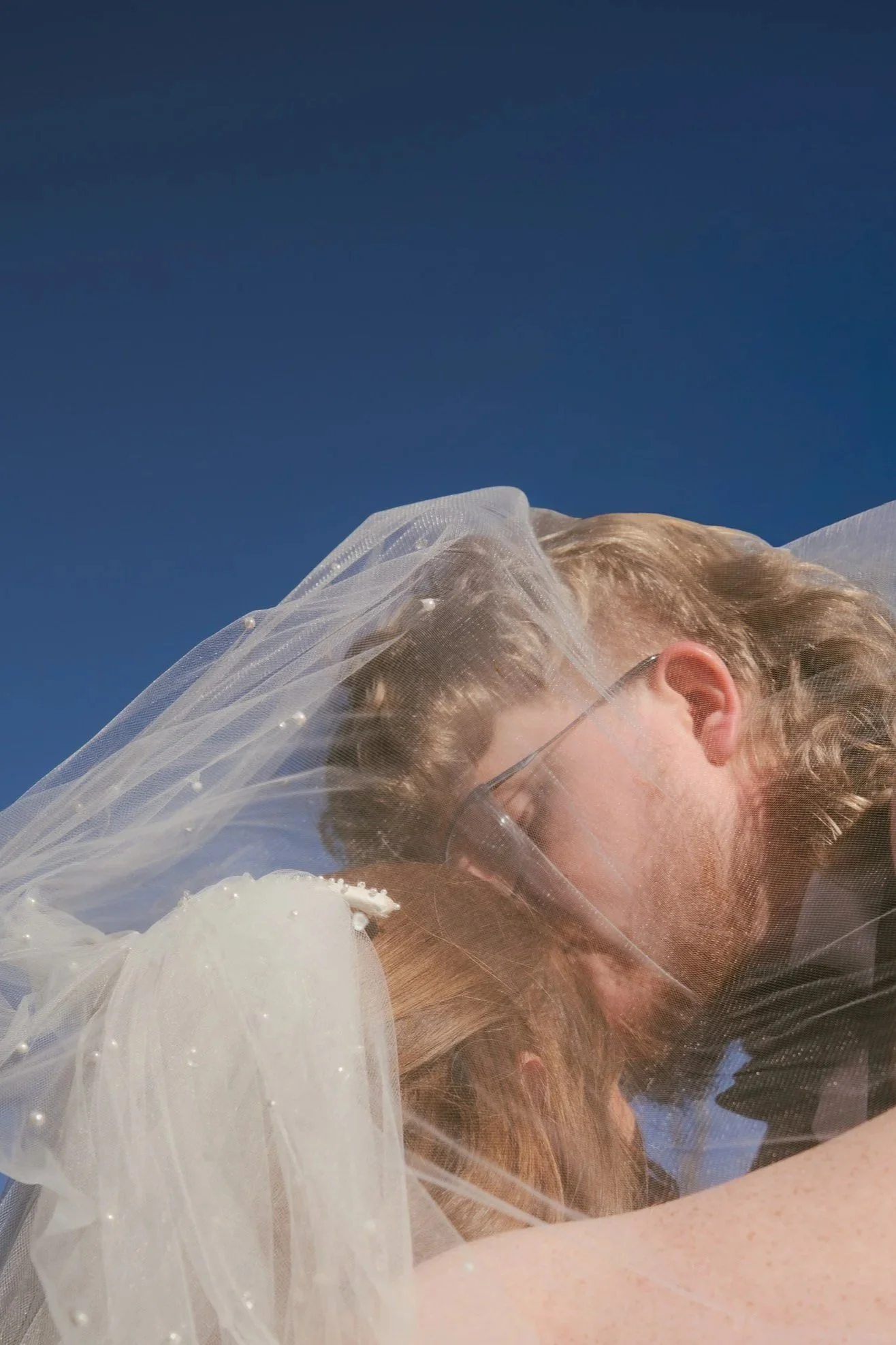 A close-up of a bride with blonde hair wearing a white wedding dress and veil, possibly outdoors against a dark blue sky.