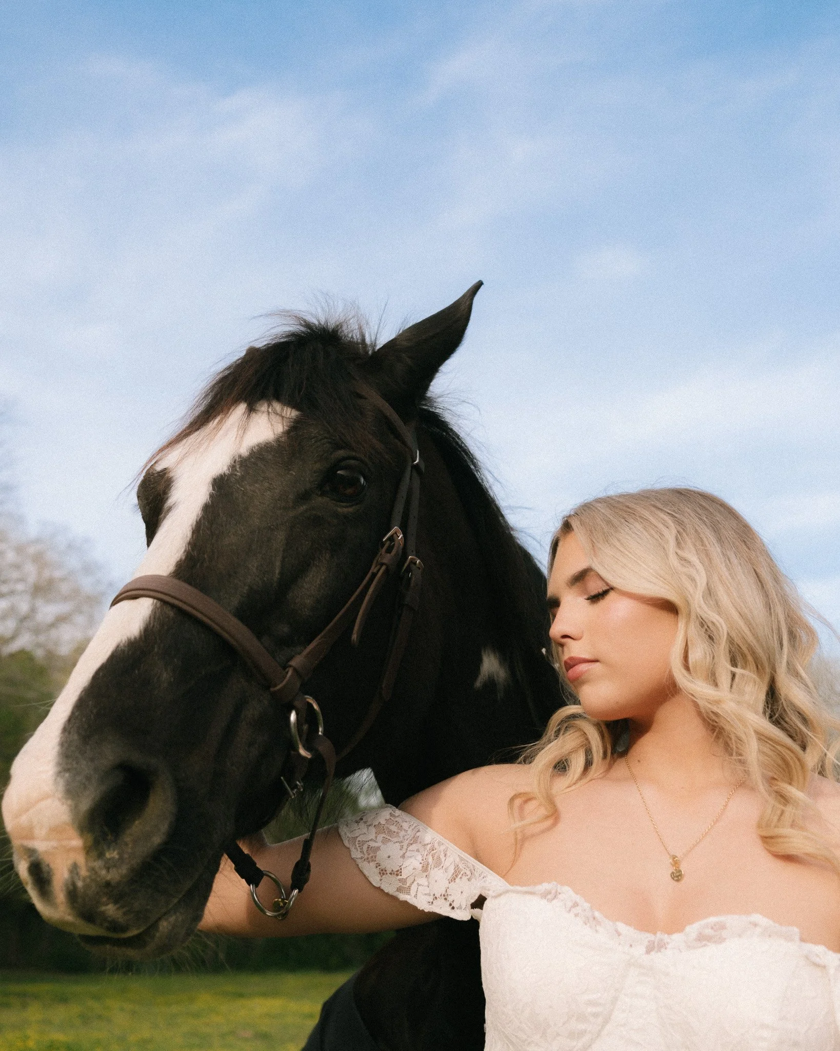 A woman with blonde curly hair and a white lace dress is close to a black and white horse, touching its face, with a blue sky and some trees in the background.