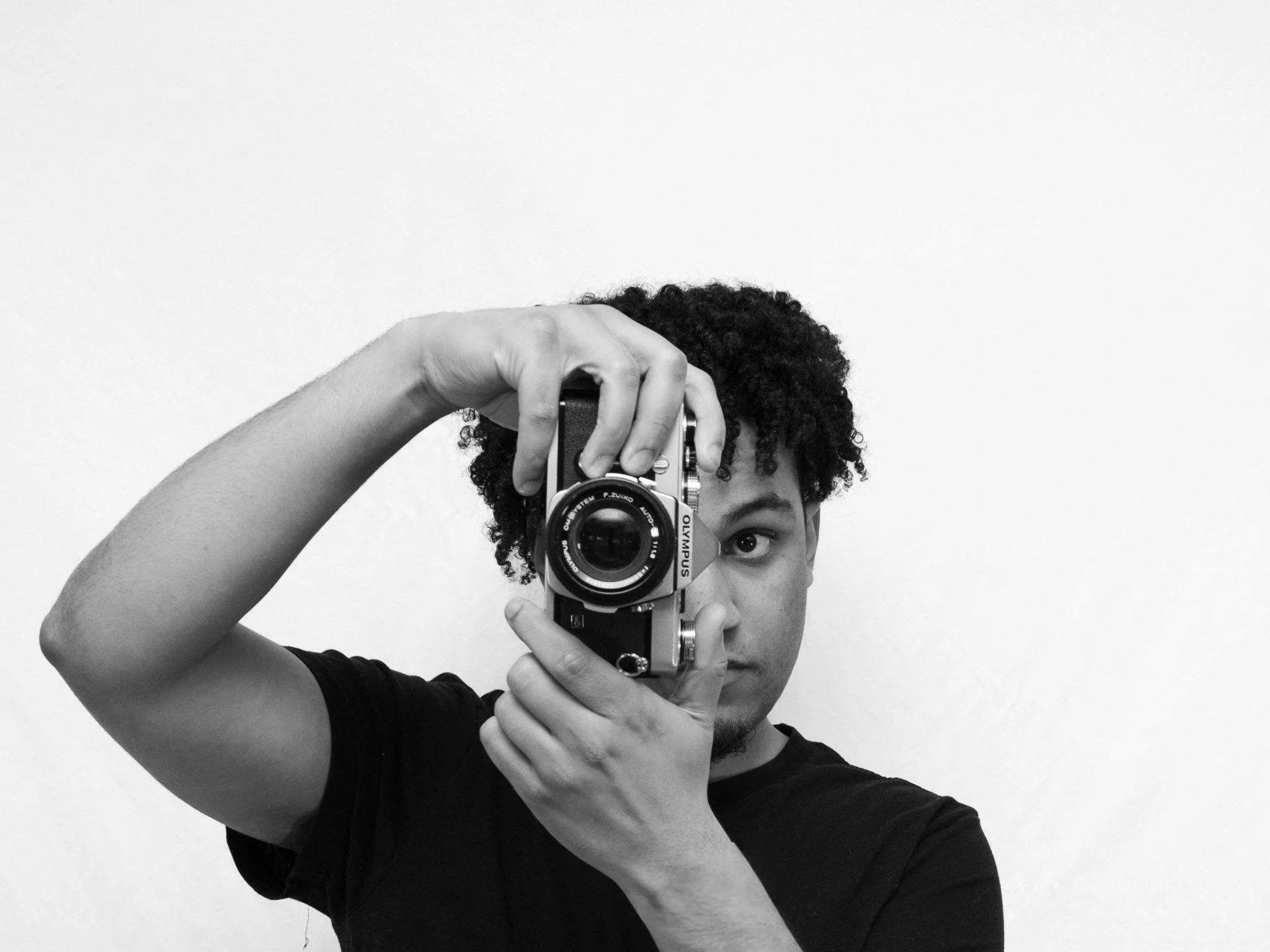 Person with curly hair taking a self-portrait with a camera, facing a white wall.
