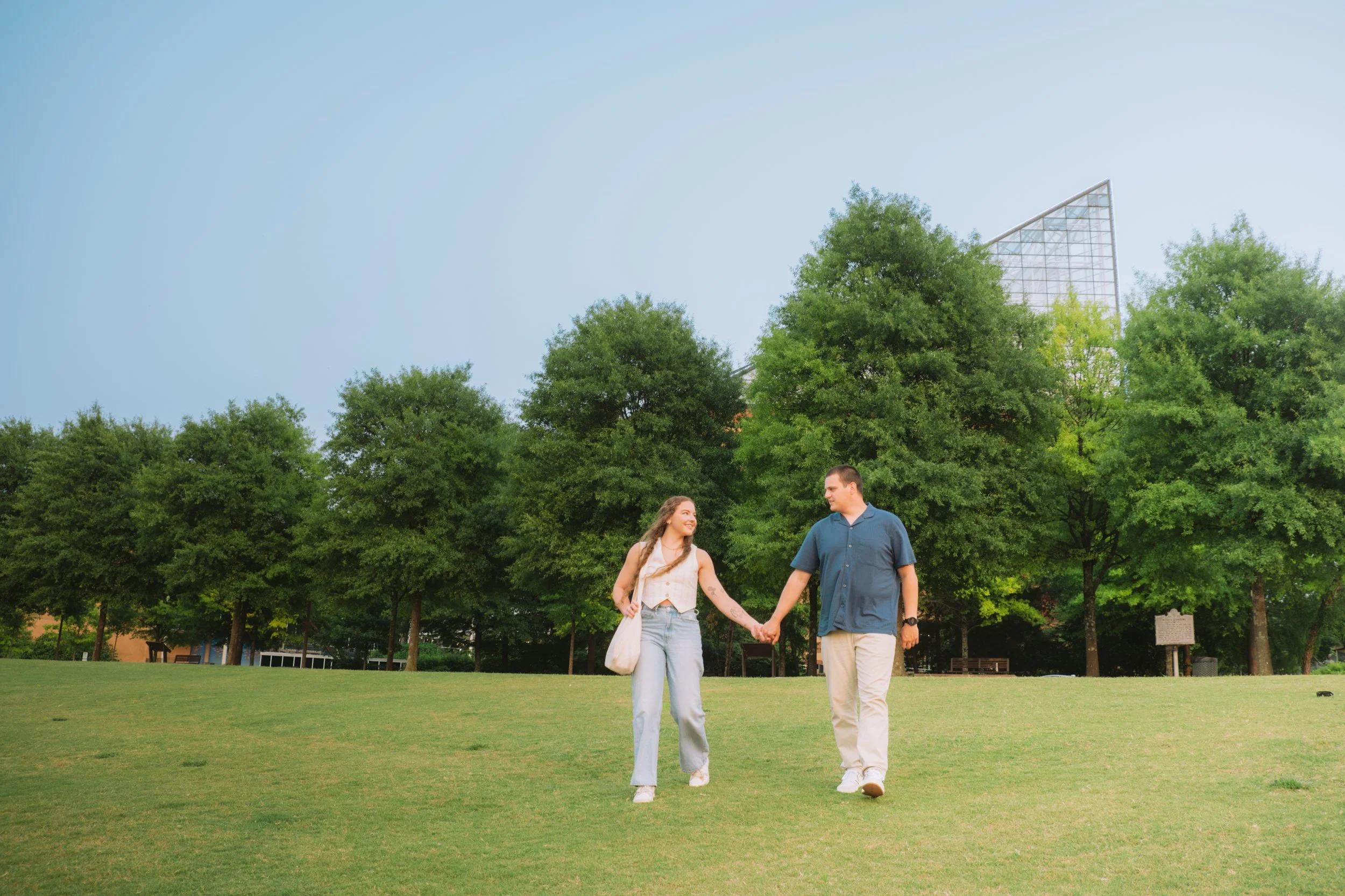 A young couple holding hands and walking together in a park with green grass and tall trees. The woman has long, curly hair, and the man has short hair and is wearing a blue shirt. They are smiling and enjoying each other's company.