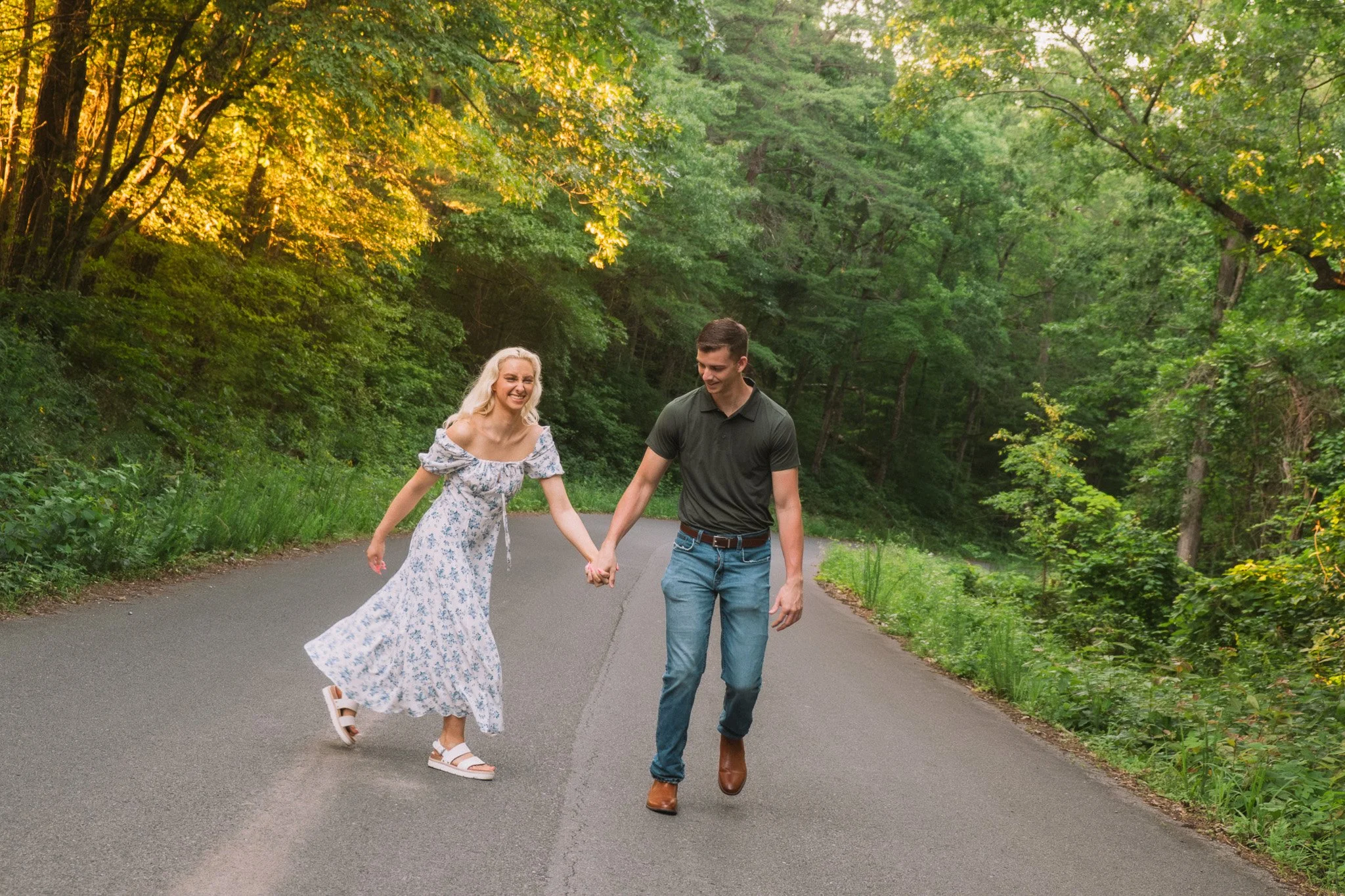 A happy couple holding hands and running on a paved forest trail during daytime, surrounded by green trees and sunlight.