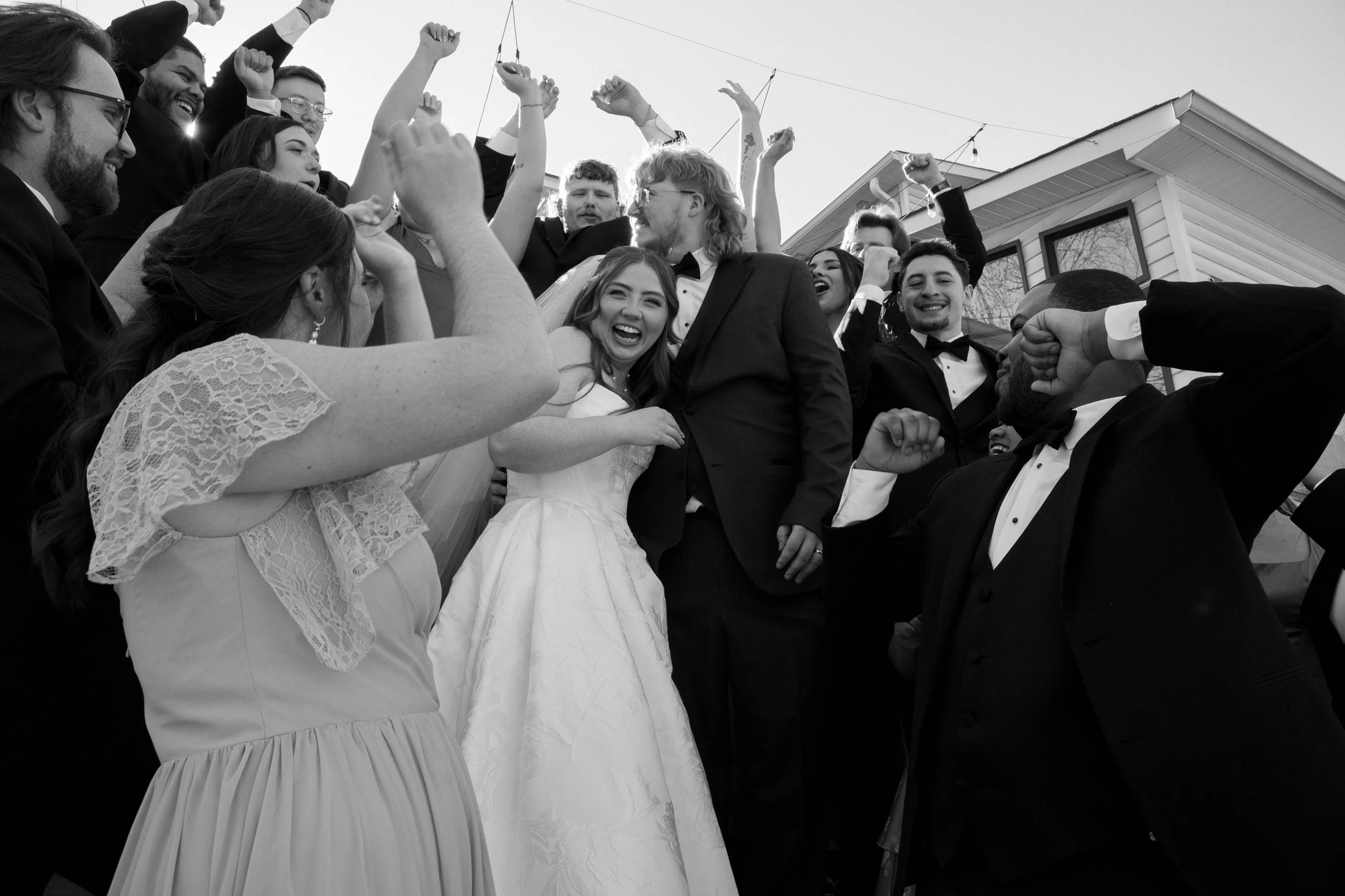 Group of people celebrating outdoors, with some raising their fists and smiling, dressed in formal attire, including tuxedos and dresses, with a house in the background.