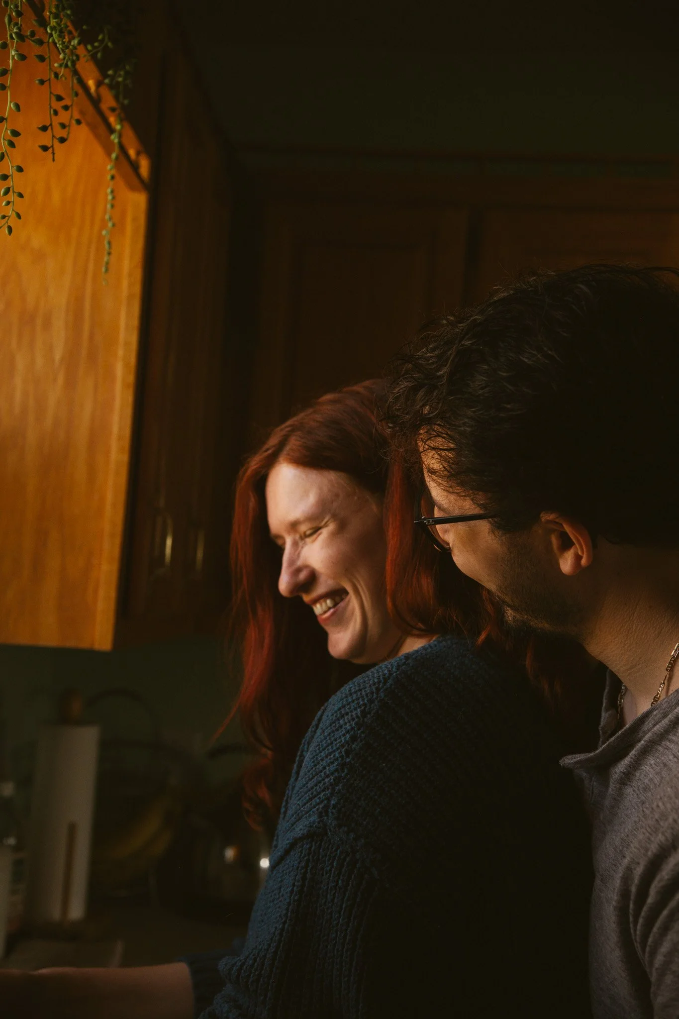 Two people sitting at a table, smiling and laughing, in a cozy, dimly lit setting with wooden walls and some greenery visible in the background.