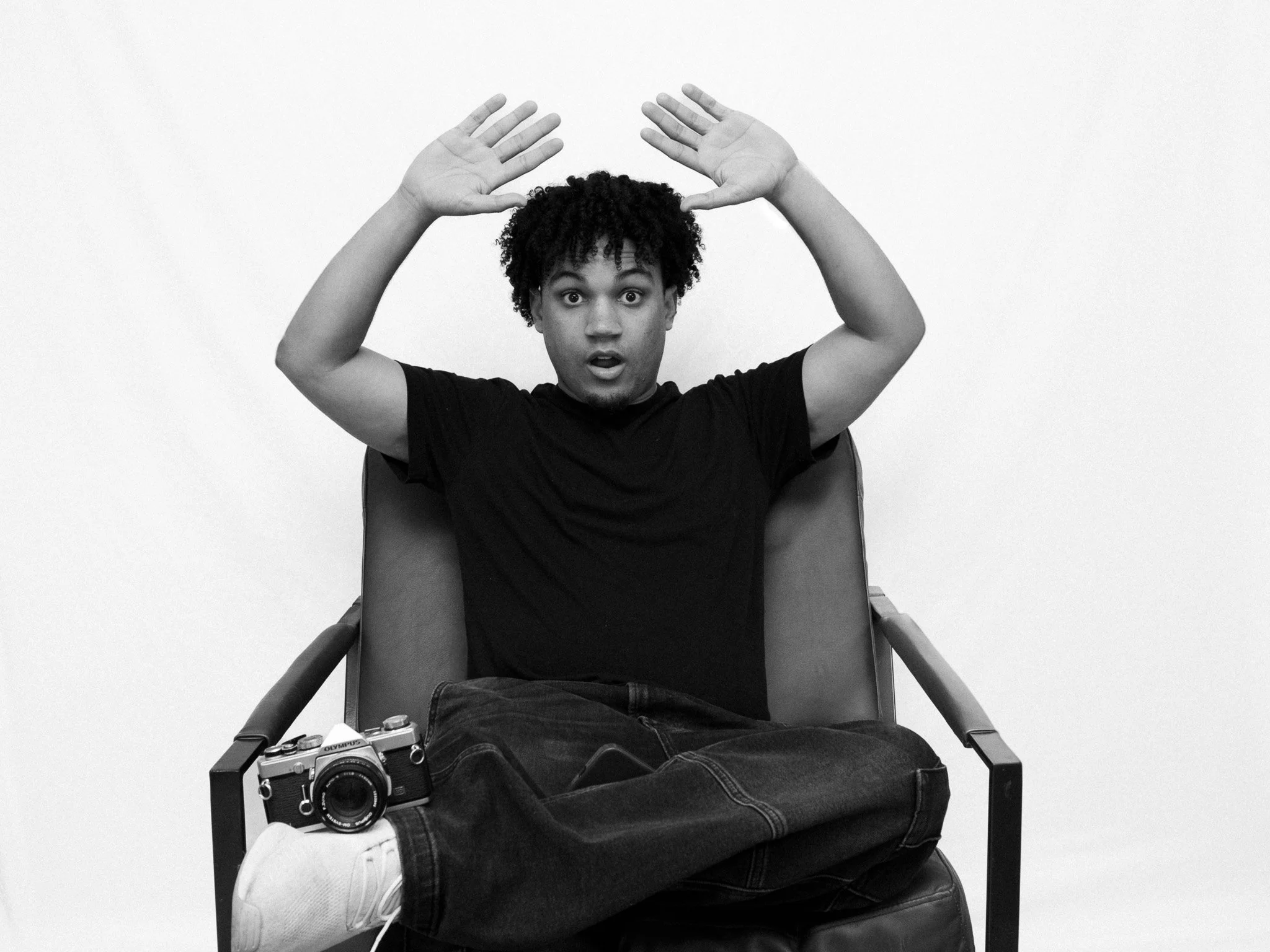 A young man with curly hair, wearing a black T-shirt and jeans, sitting in a chair with his hands raised above his head in front of a plain white wall, with a camera on his lap.