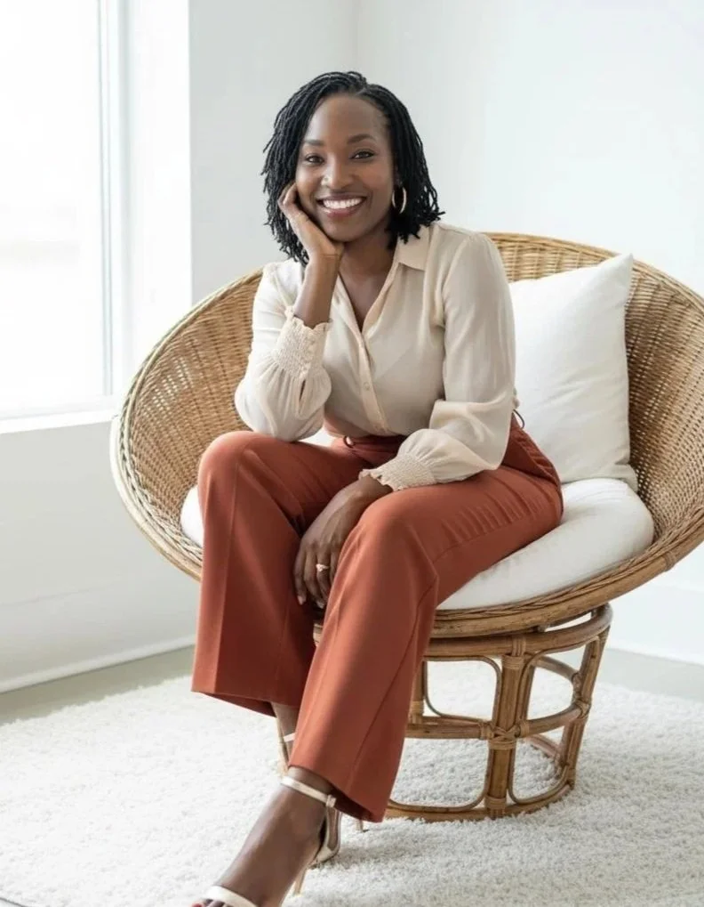 A woman with shoulder-length dreadlocks sitting on a wicker chair, smiling, in a well-lit room with a white wall and a white pillow behind her.