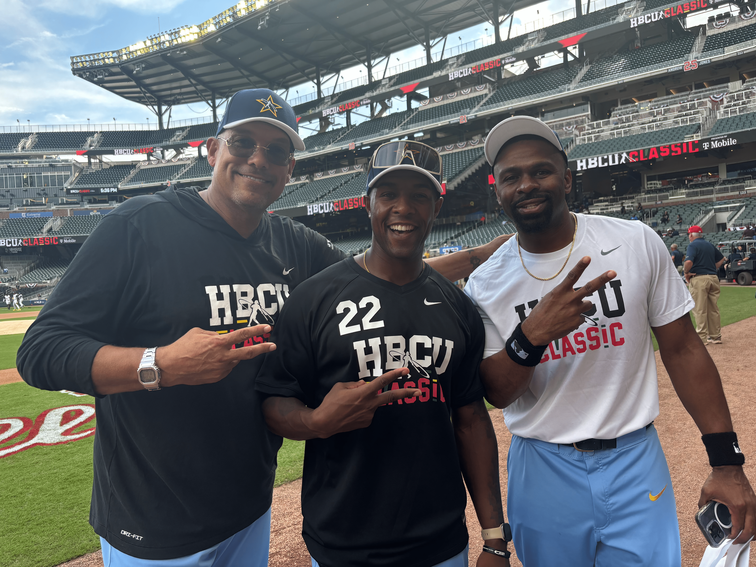 Three men smiling and posing at a baseball stadium, two wearing black HBCU Classic shirts and one in a white shirt. They are making peace signs with their hands. The stadium is filled with empty seats and a large scoreboard in the background.