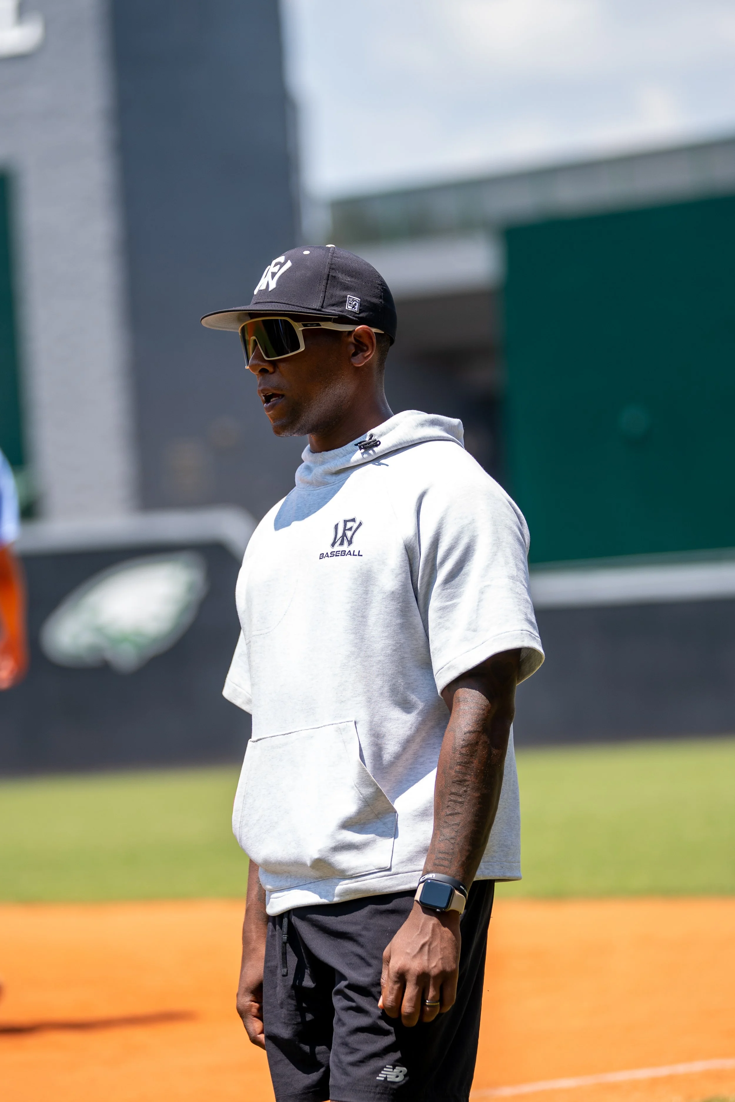 Man wearing a black cap, sunglasses, a light grey hoodie with 'LF Baseball' logo, black shorts, and a smartwatch standing on a baseball field.