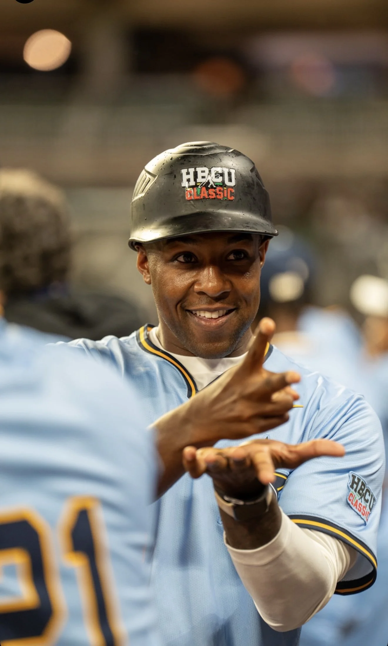 Young man in baseball uniform smiling and pointing, wearing a black helmet with 'HBO Classic' logo, at a baseball game.