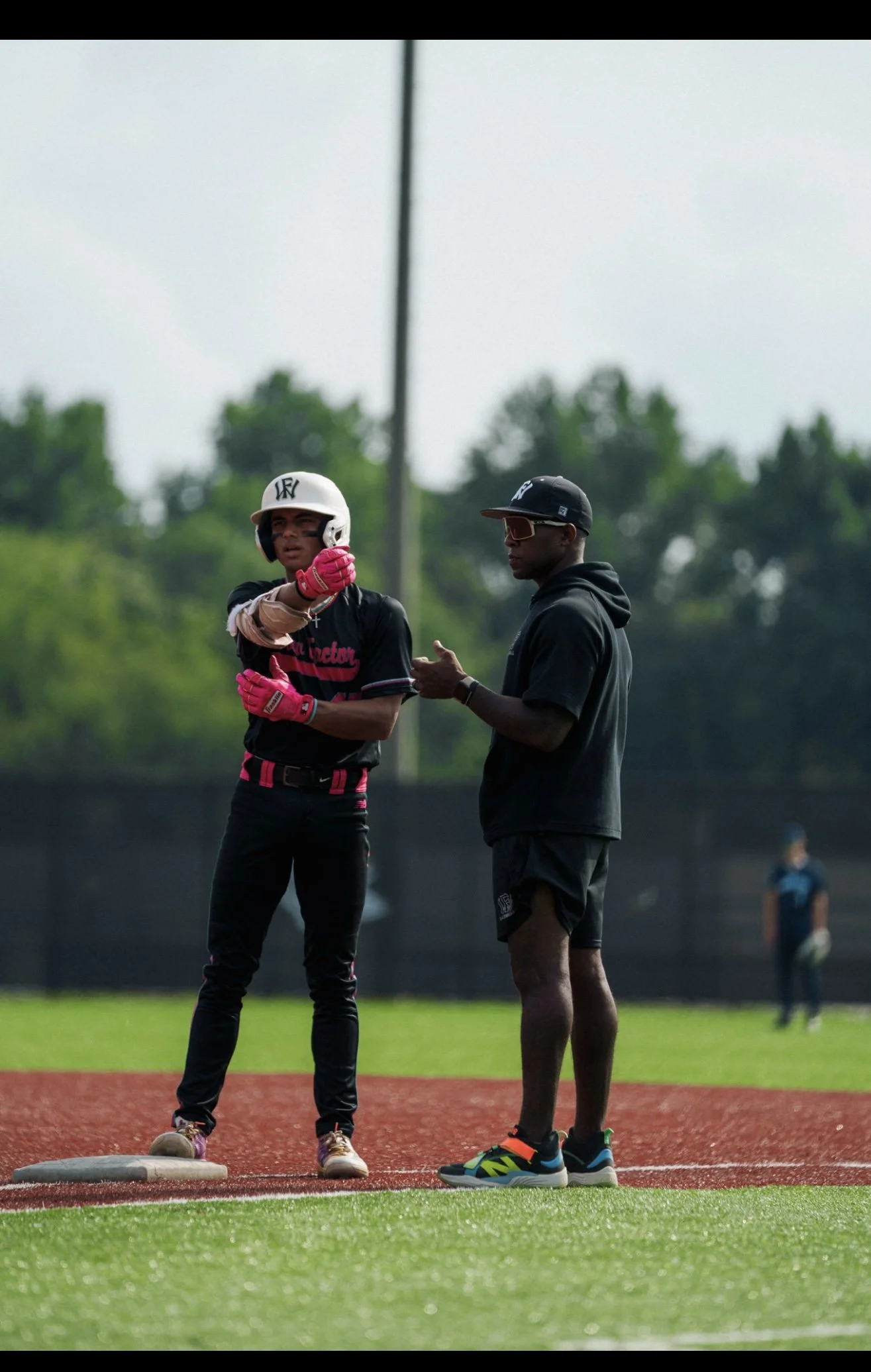 A young male baseball player in a black uniform with pink gloves and a white helmet standing near a base on a baseball field, talking to a coach in black clothing with sunglasses, wearing sneakers, as they discuss during a game.