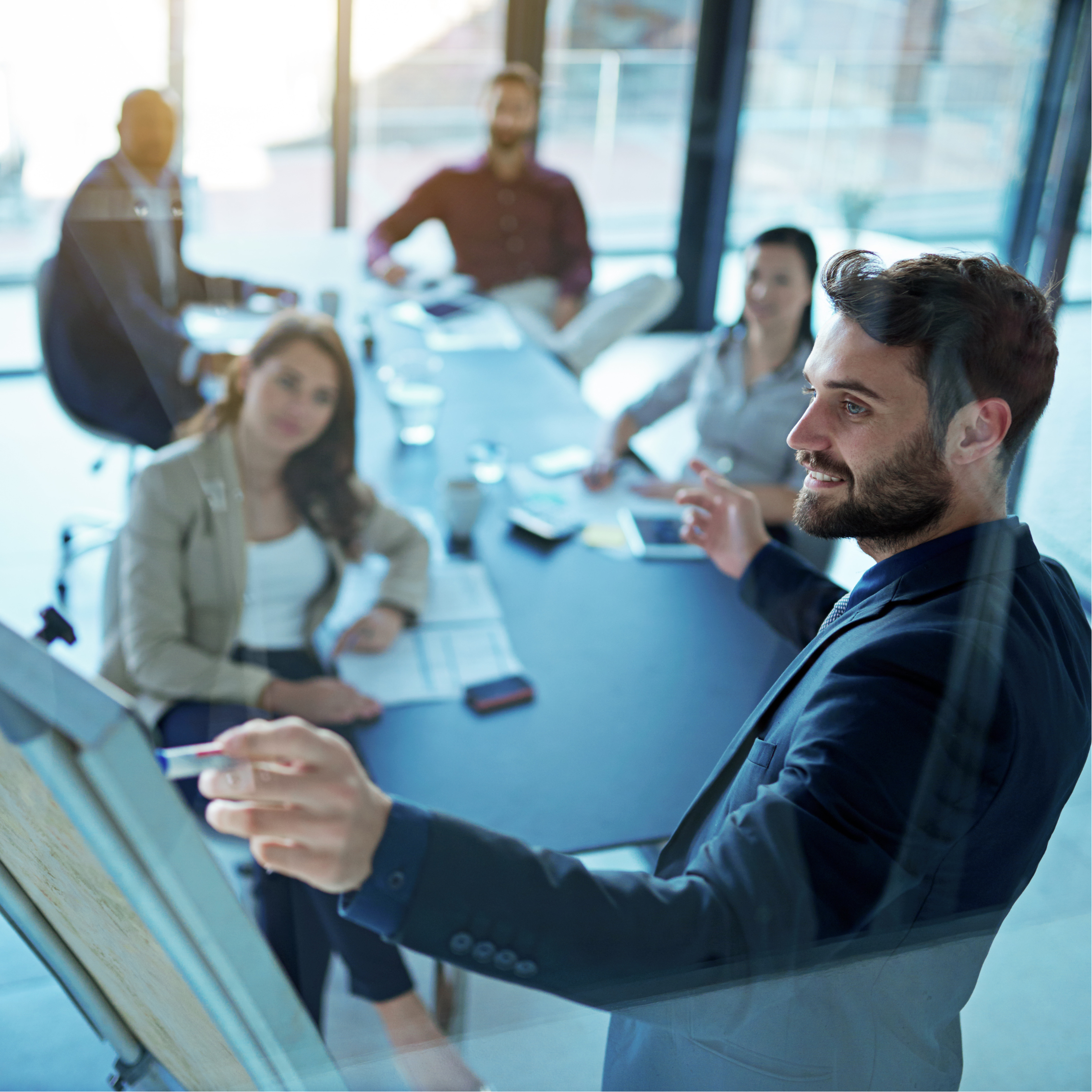 man pointing to a presentation board in front of four other professionals around a board table in front of windows