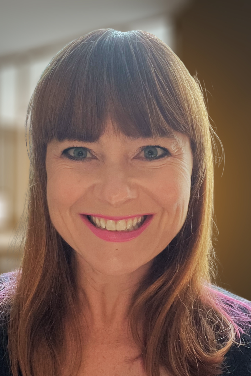 Smiling woman with shoulder-length light brown hair, blue eyes, wearing a black blazer and a red top, in front of a brick wall background.