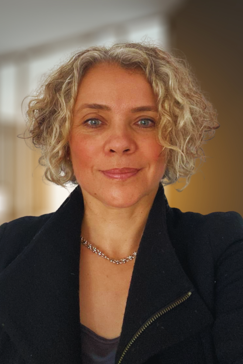 A woman with curly blonde hair, wearing a green sweater, blue jeans, a scarf, and an Apple Watch, smiling while standing in a brightly lit, modern hallway with plants on the windowsill.