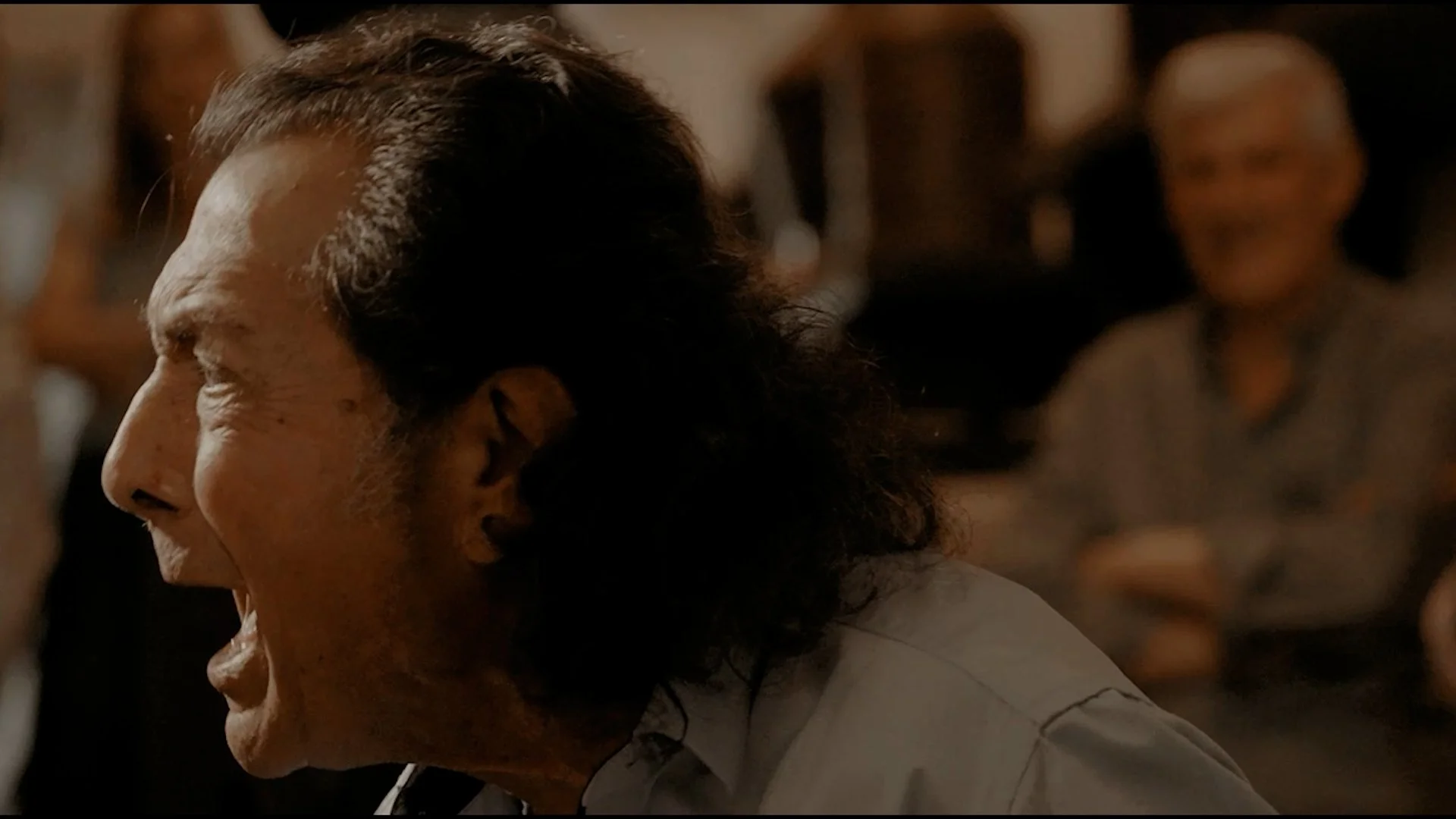 Hombre de perfil con cabello rizado, rostro en silencio, en un ambiente con otras personas en el fondo.