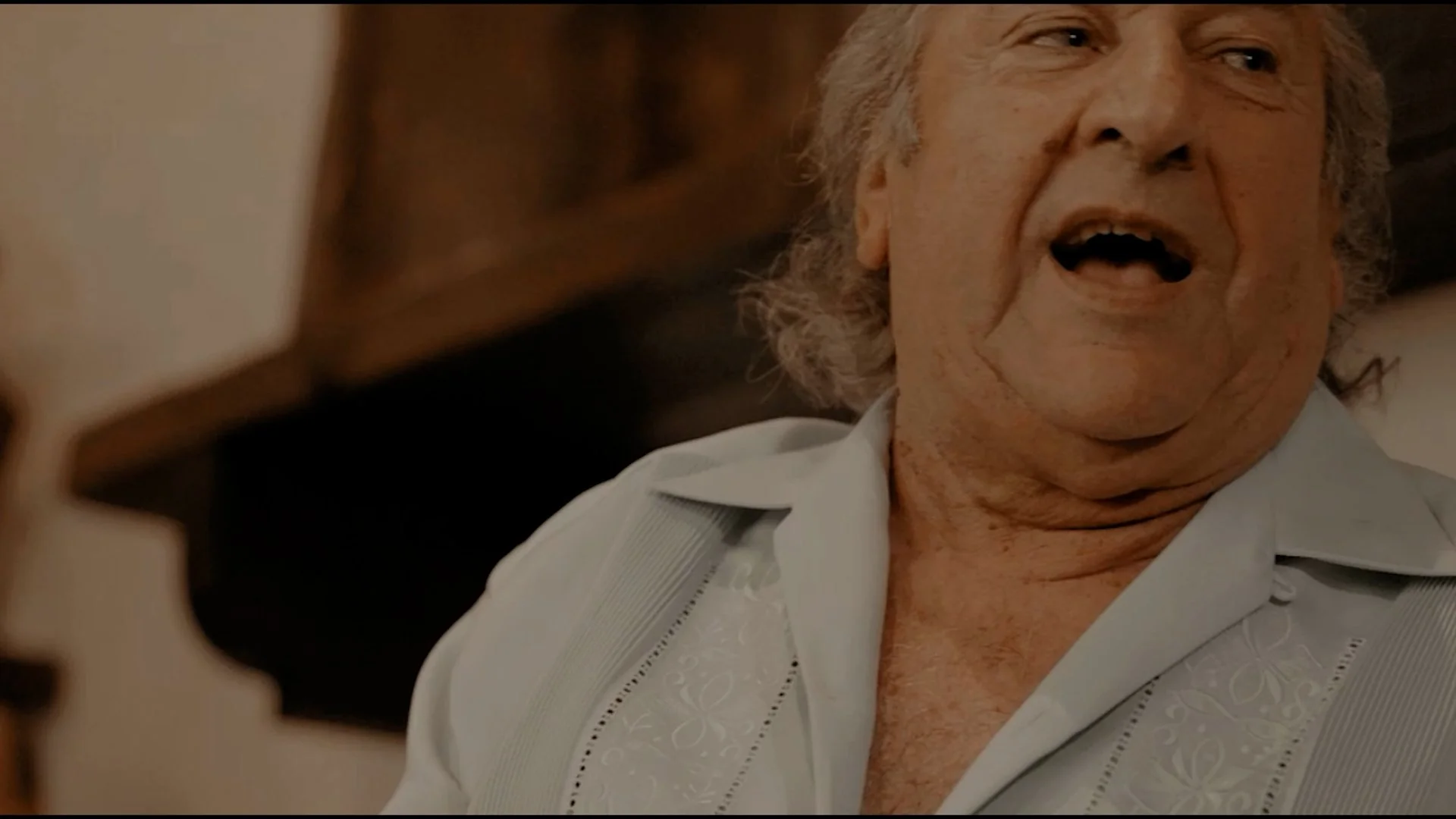 Anciano con cabello gris y expresión feliz, usando una camisa blanca con bordados, sentado en un ambiente interior.