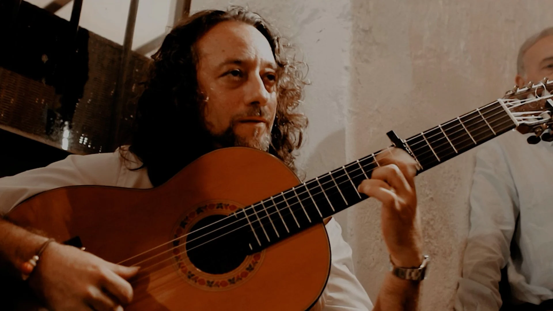 Hombre tocando guitarra acústica, con cabello rizado y barba, en un ambiente interior con paredes de piedra.