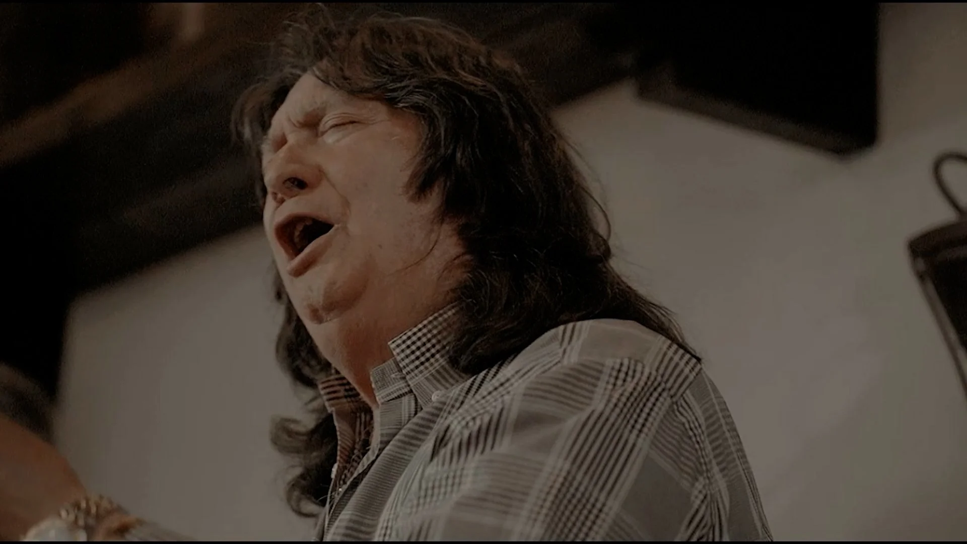 Hombre con cabello largo y rizado en un momento de canto, con expresión v estremecimiento, usando camisa de cuadros, en un entorno interior.