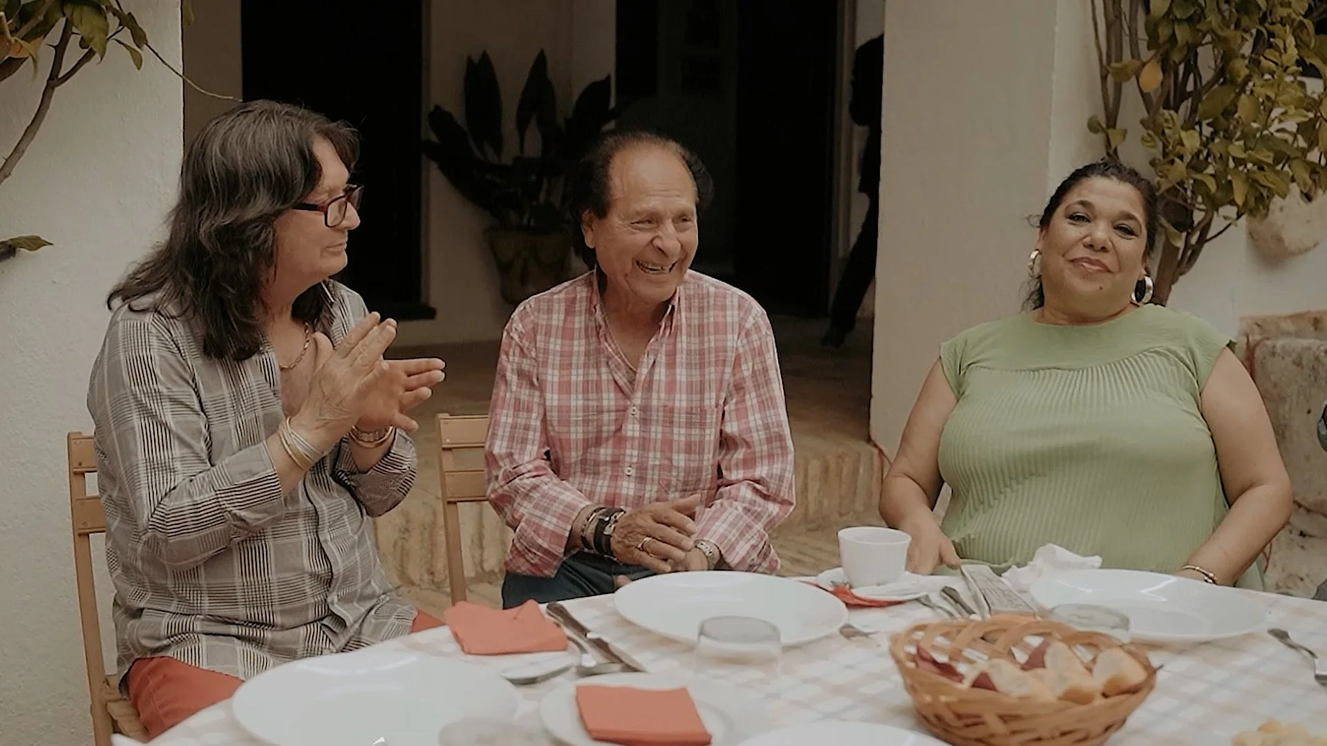 Tres personas sentadas en una mesa conversando y sonriendo en un ambiente acogedor, con platos y utensilios en la mesa.