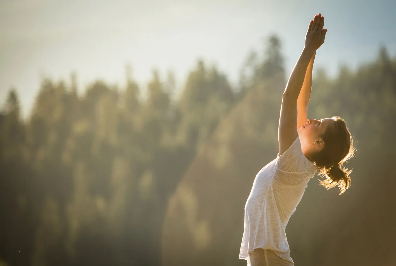 A woman in a white t-shirt stands outdoors with her arms raised above her head, reaching toward the sky, in a natural setting during what appears to be sunrise or sunset.