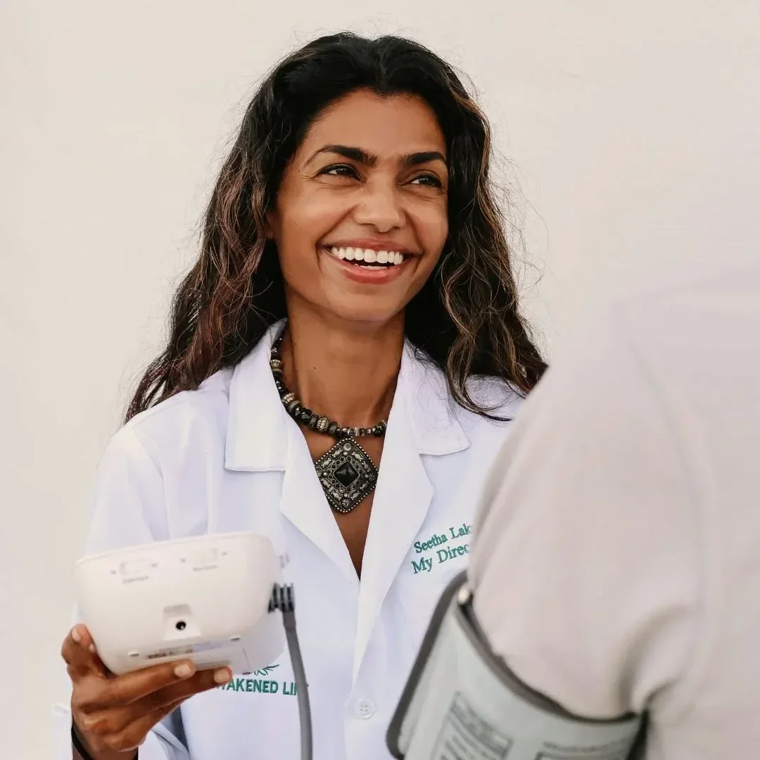 Dr. Seetha Lakshmi wearing a white lab coat, holding medical equipment, smiling and talking to a healthcare professional.