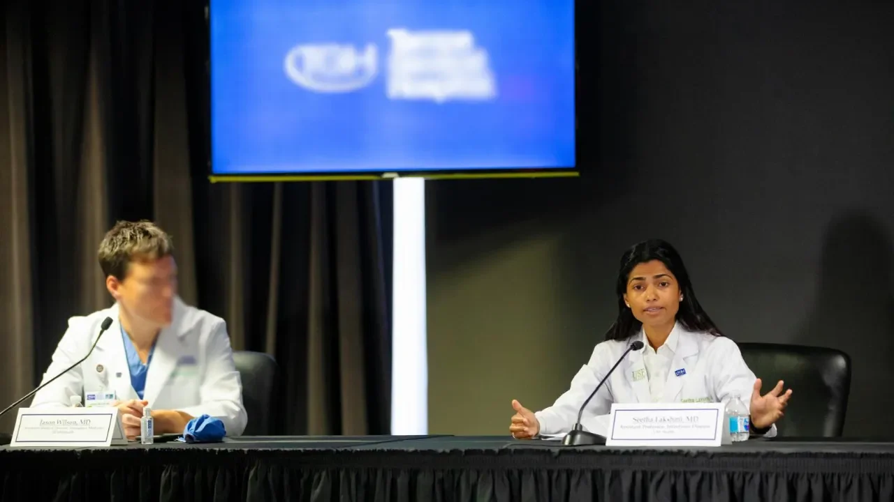 Two female doctors in white lab coats are speaking at a panel discussion at a conference. One is gesturing with her hands, and the other has blurred facial features. A large screen displaying the Intel logo is in the background.