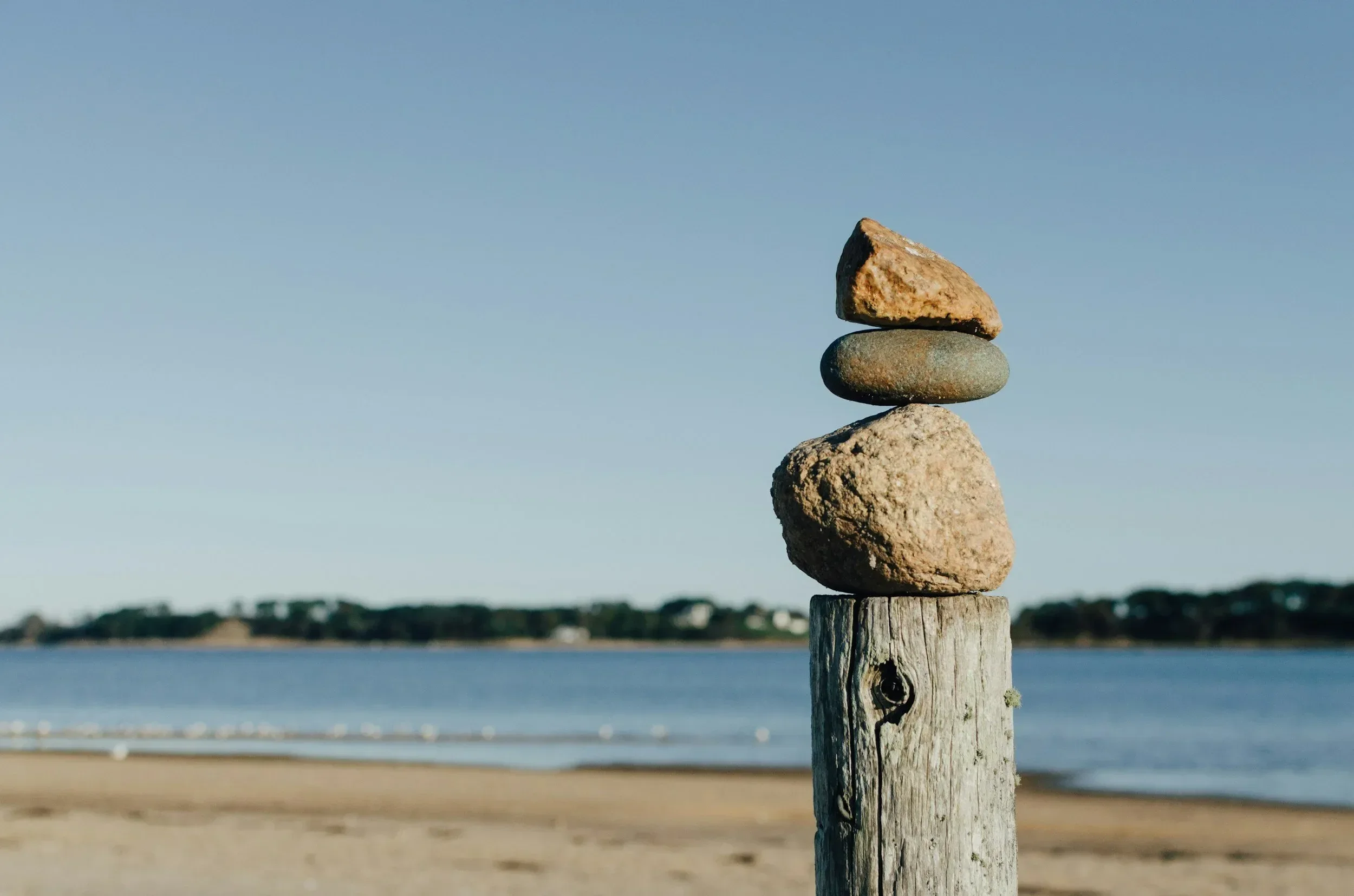Stacked rocks on a wooden post by a beach, with water and trees in the background under a clear sky.