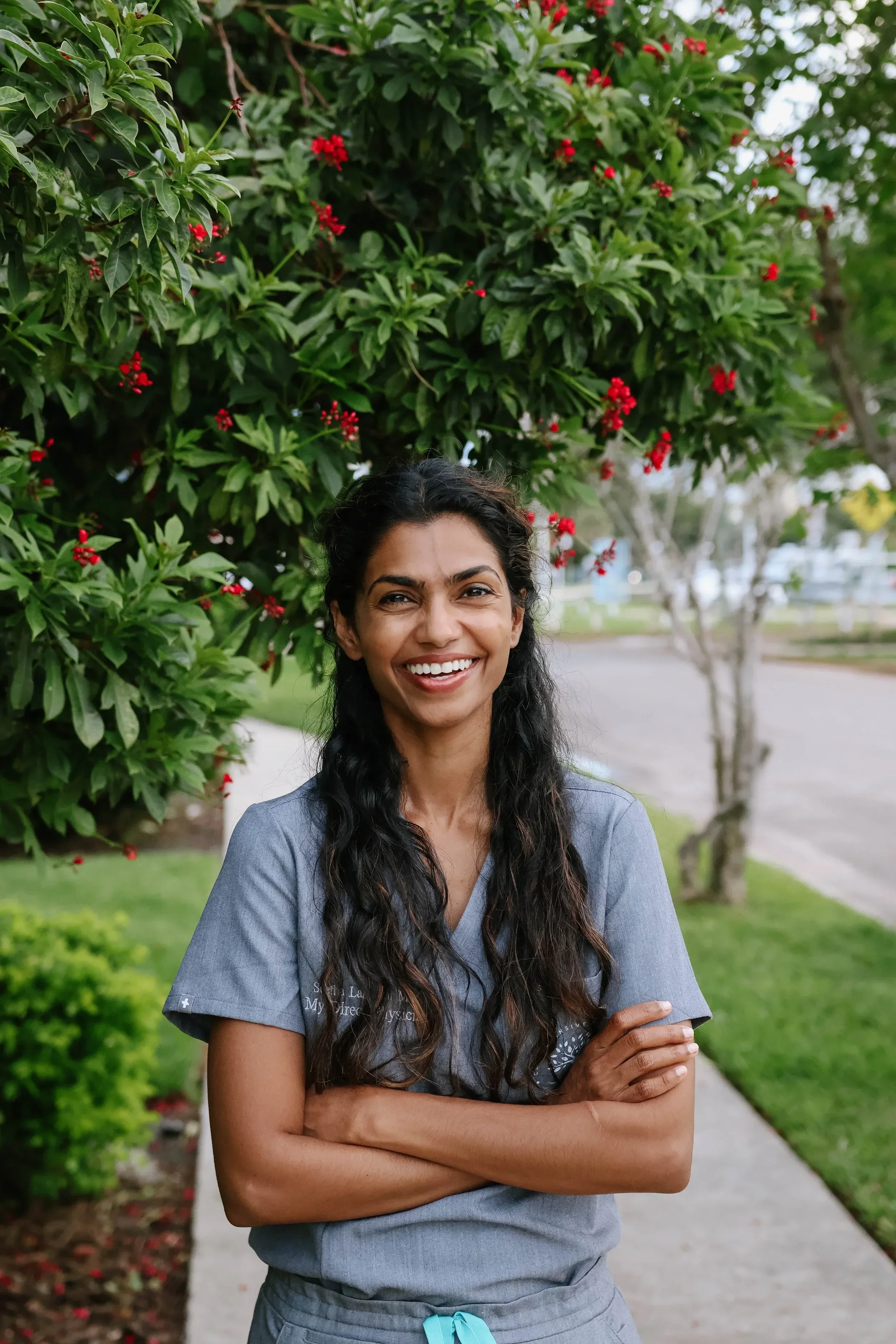 Dr. Seetha Lakshmi with long curly dark hair standing outdoors in front of a bush with red flowers and green leaves, smiling with her arms crossed, wearing a gray medical scrub top with white text and symbols, and gray pants with a blue drawstring.