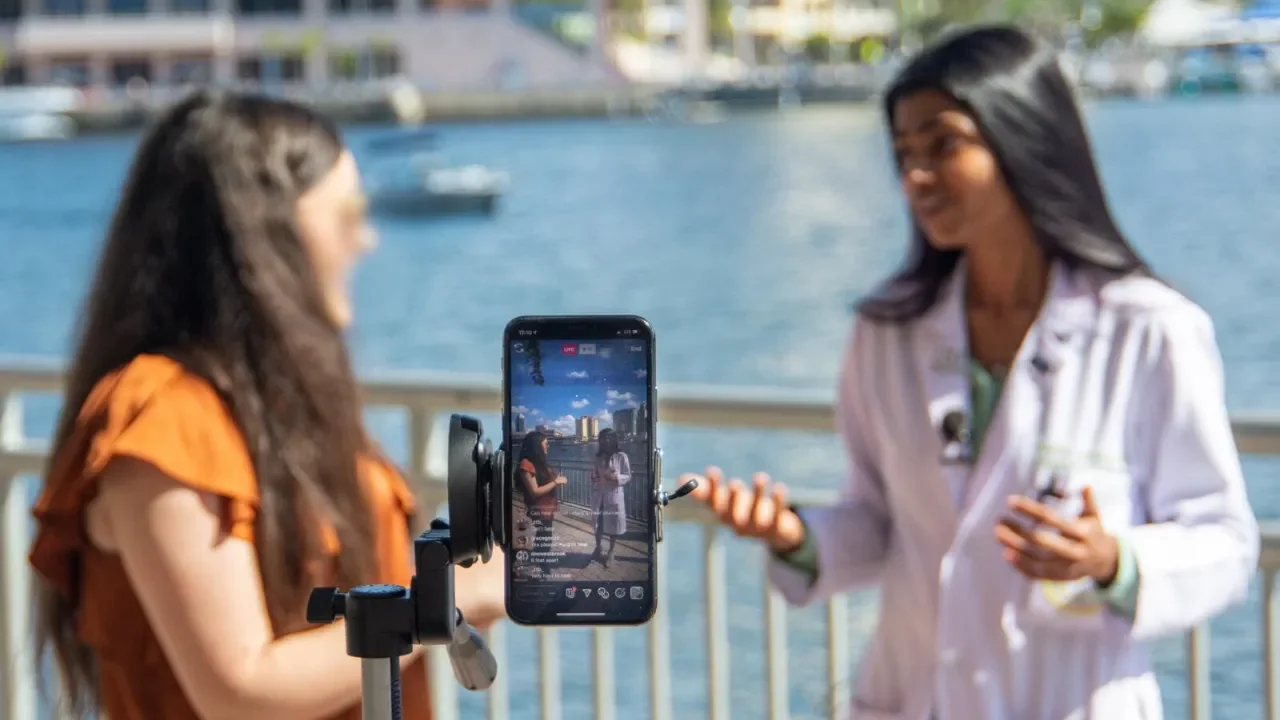 Dr. Seetha Lakshmi and a woman having a conversation outdoors by the water, with one holding a microphone and the other speaking in front of a camera mounted on a tripod, during daytime.