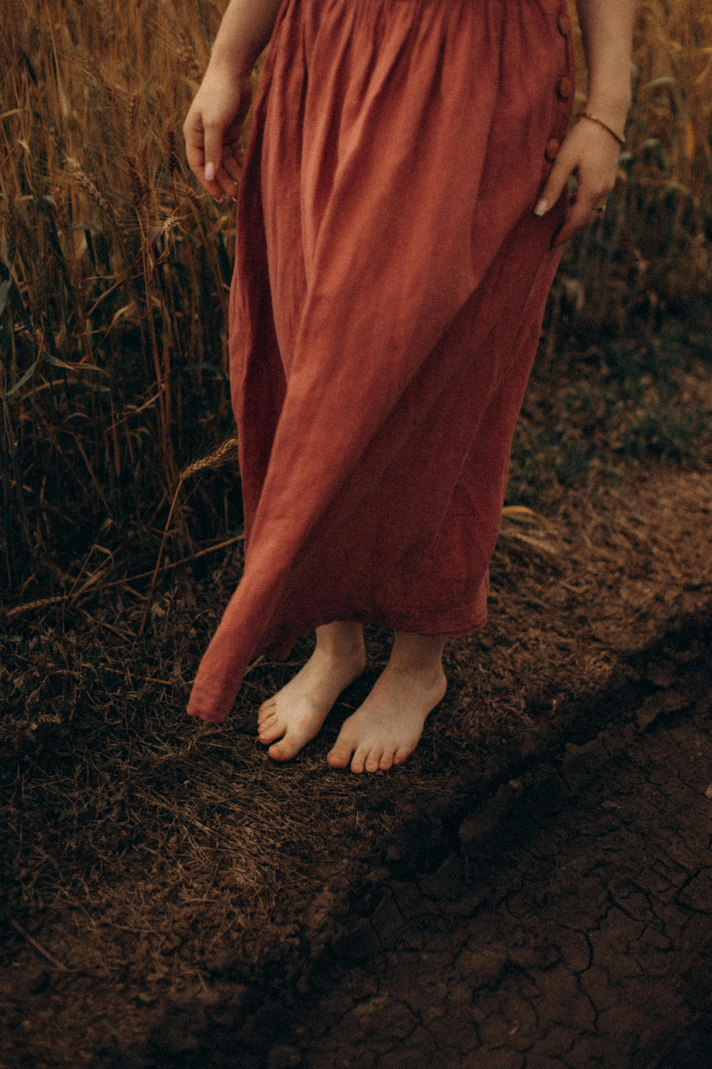 Person in a long rust-colored skirt standing barefoot on cracked dry ground amidst tall dry grass.
