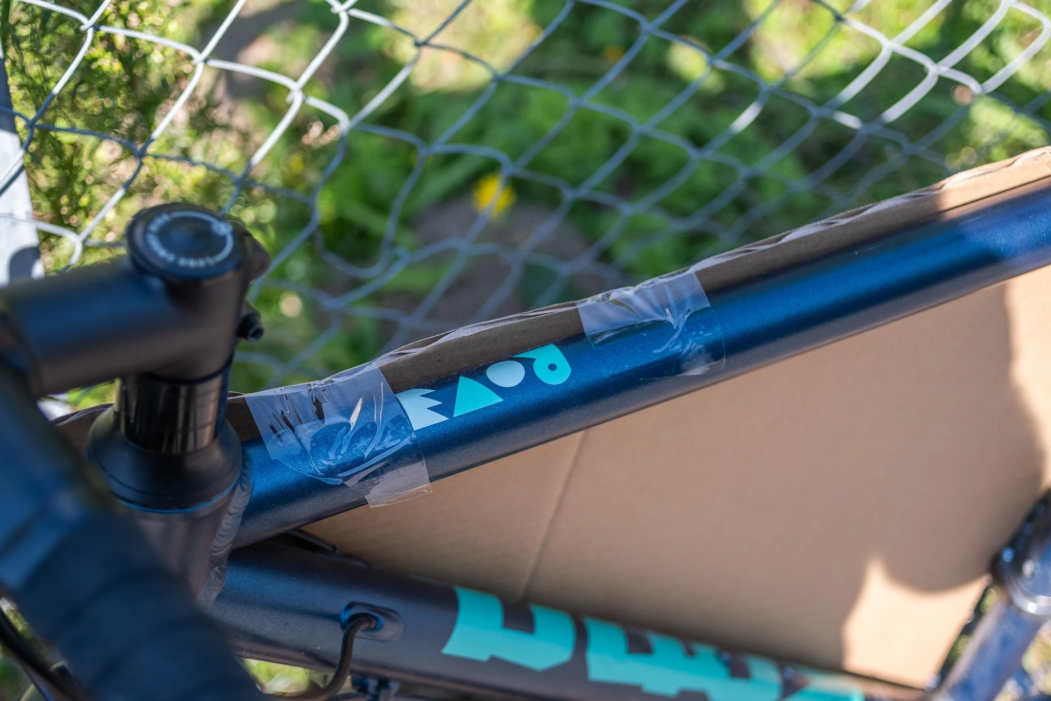 Un cadre de vélo avec une tige de direction, une selle et une carte en carton attachée avec du ruban adhésif, à côté d'une clôture en métal et de végétation.