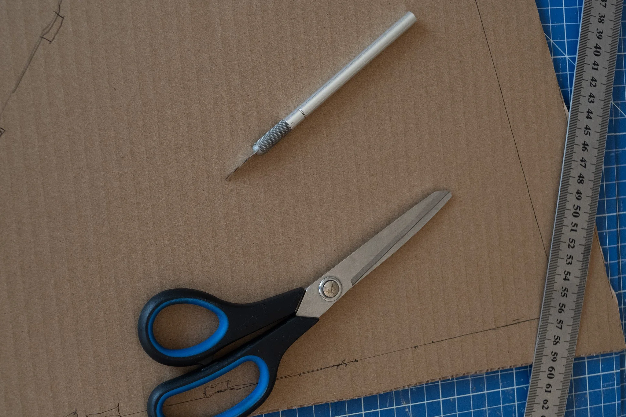 Une paire de ciseaux, un cutter, une règle métallique, et un morceau de carton avec des lignes dessinées, utilisés pour la fabrication ou le bricolage.