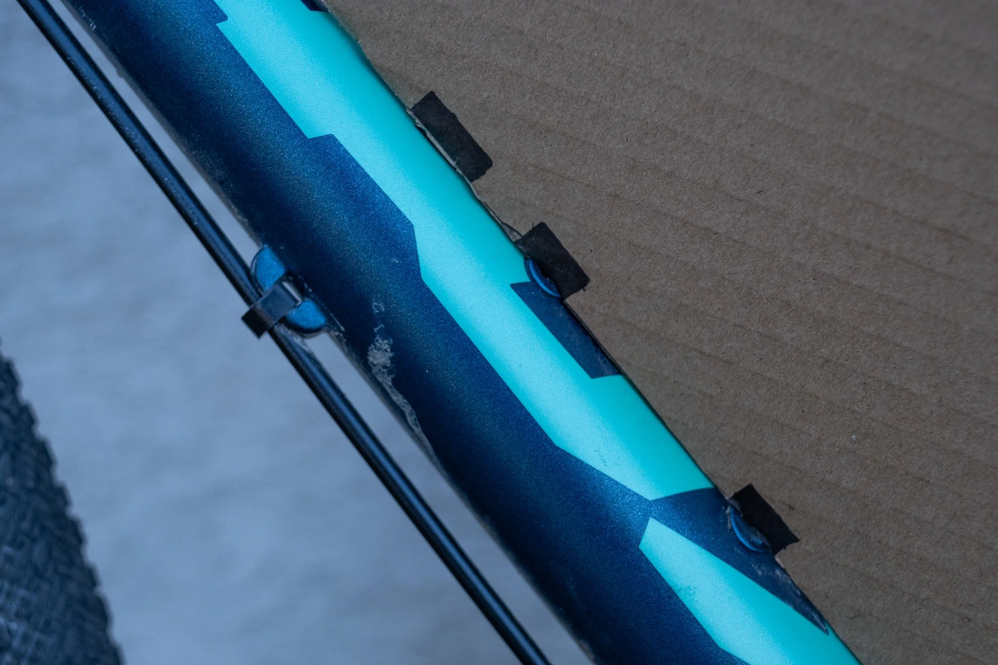 Zoom sur une partie d'un vélo ou d'un matériel sportif, de couleur noire avec des accents bleus, fixé à un fond en carton ou en matière similaire.