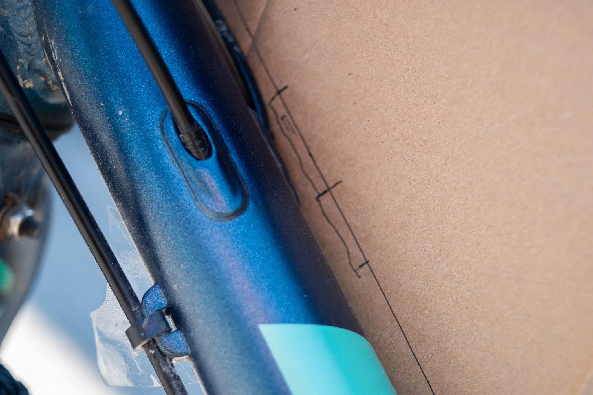Détail d'un cadre de vélo en métal bleu, fixé à une surface en carton ou en bois, avec un fil électrique noir passant à travers une ouverture dans le cadre.