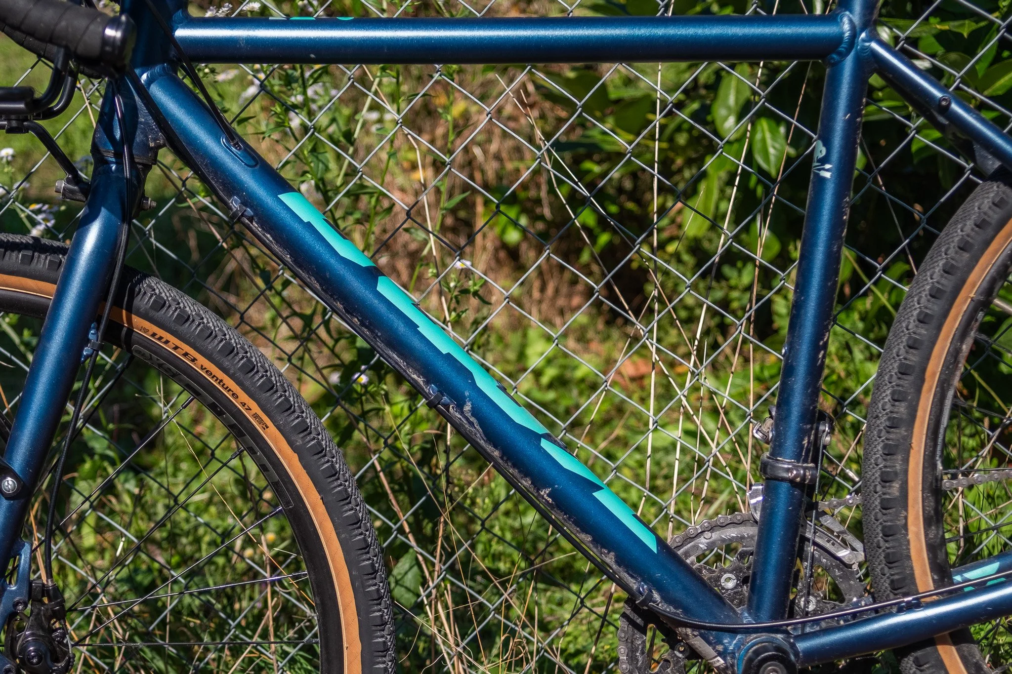Vélo bleu garé contre une clôture en fil métallique, avec des pneus à flancs beiges et un terrain herbeux en arrière-plan.
