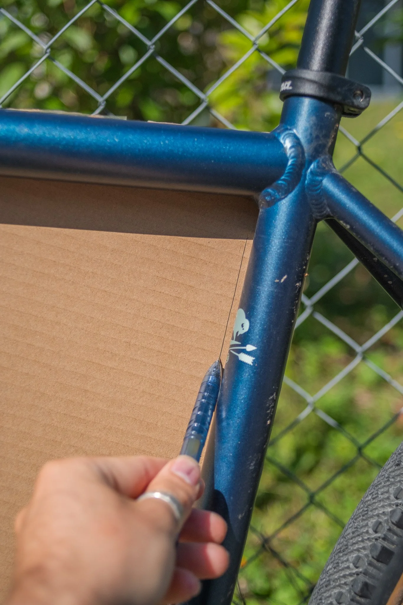 Une main avec une bague tire une aiguille à une chambre à air de vélo bleue, près d'une clôture en métal et d'un panneau en carton.
