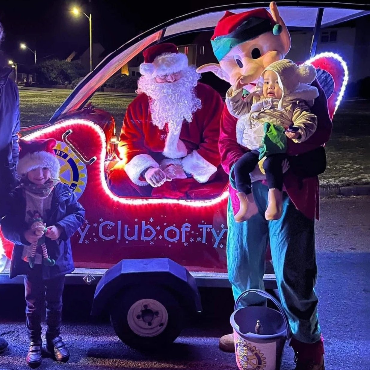 A child wearing a blue jacket and striped shoes is standing in front of a float with Santa Claus. Santa is sitting in a red sleigh, and a person dressed as an elf is holding a young child in front of the sleigh. The elf is wearing a costume with a large elf head, a red outfit, and is carrying a bucket. It is nighttime, and the scene is decorated with bright lights.