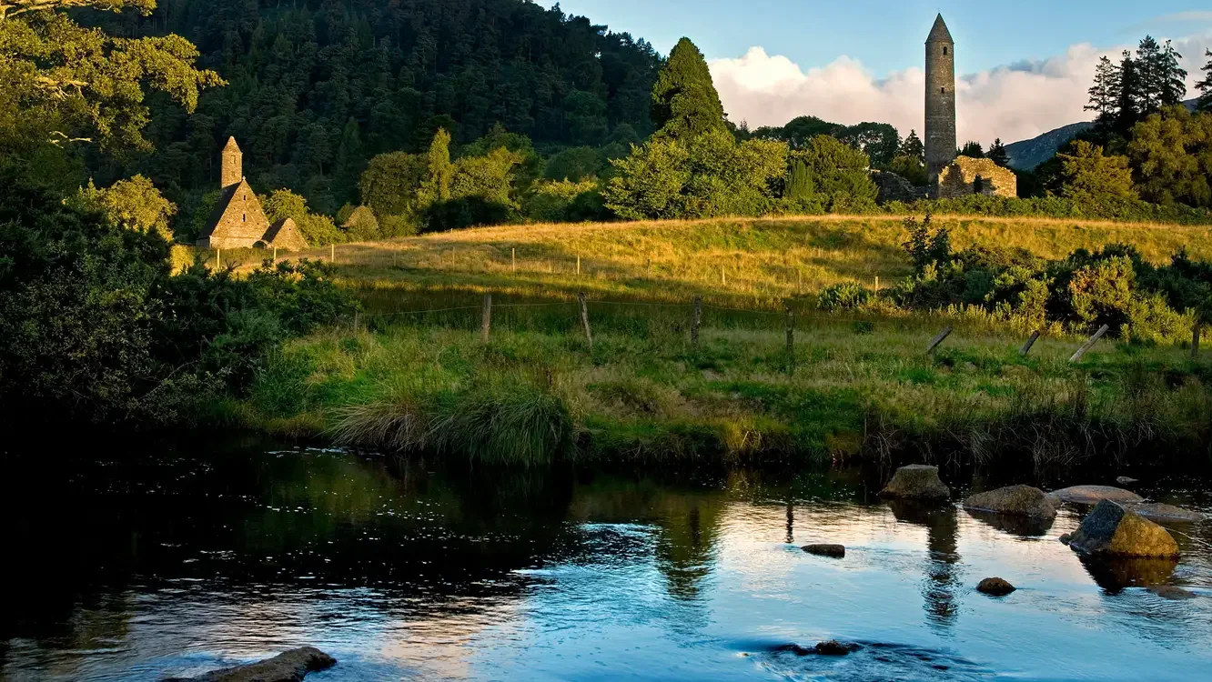 A scenic landscape featuring a river in the foreground, a grassy hillside with trees, and historic stone buildings including a tower in the background, illuminated by warm sunlight.