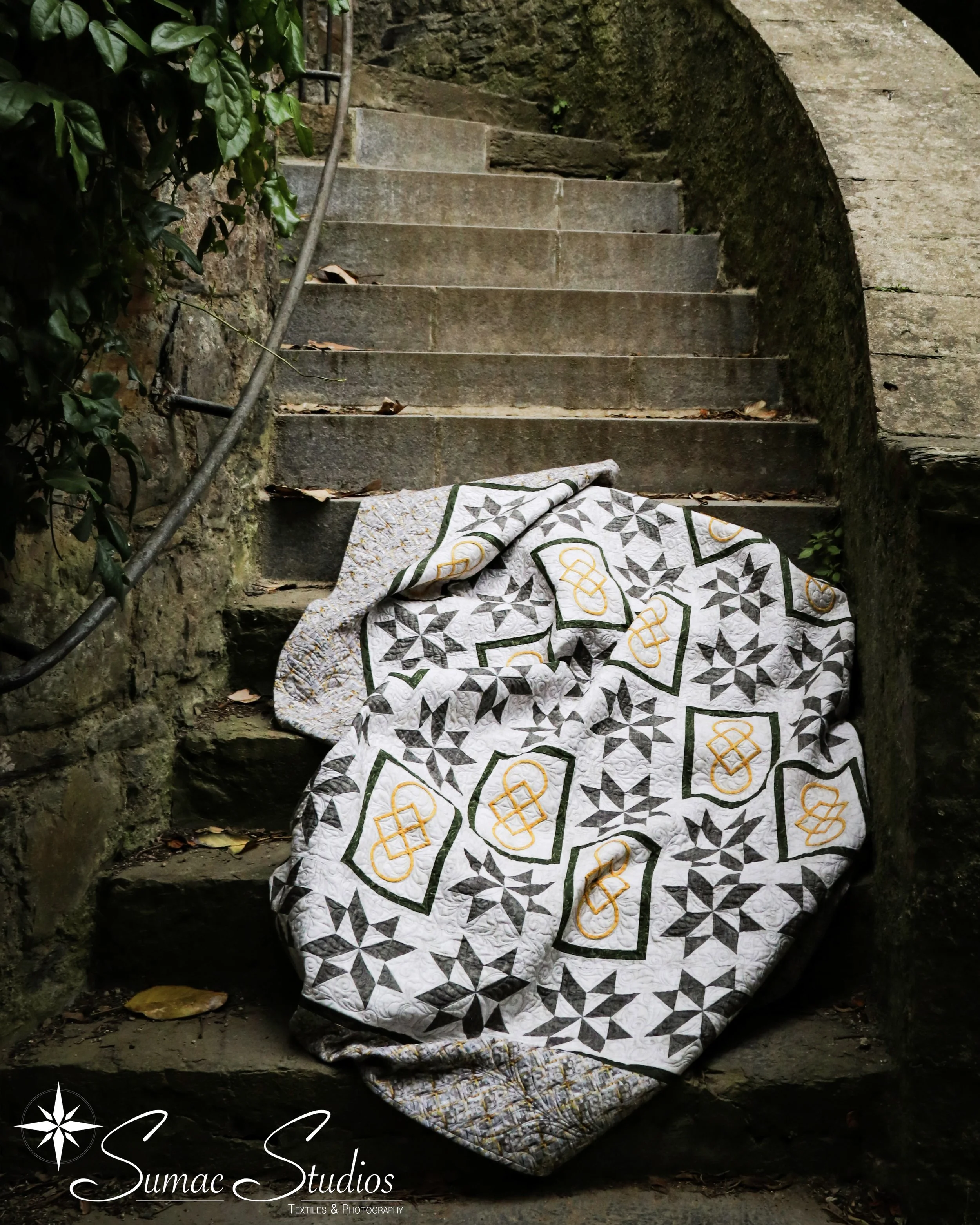 A quilt with black, white, green, and yellow patterns draped on an outdoor stone staircase with moss and fallen leaves.