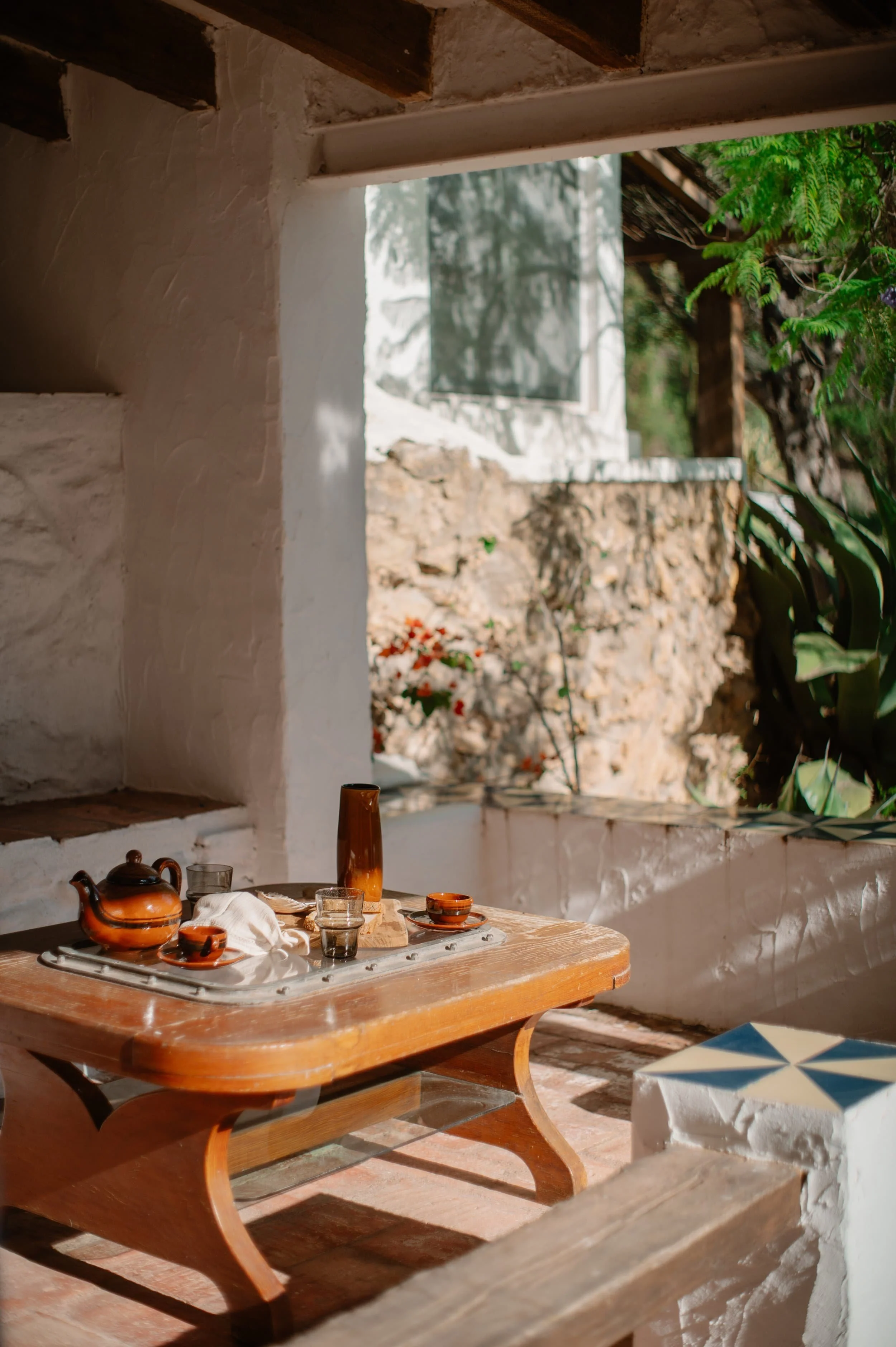 A rustic wooden table set for tea with teapot, cups, glasses, and a decorative vase outside a whitewashed building with stone walls and lush green plants.