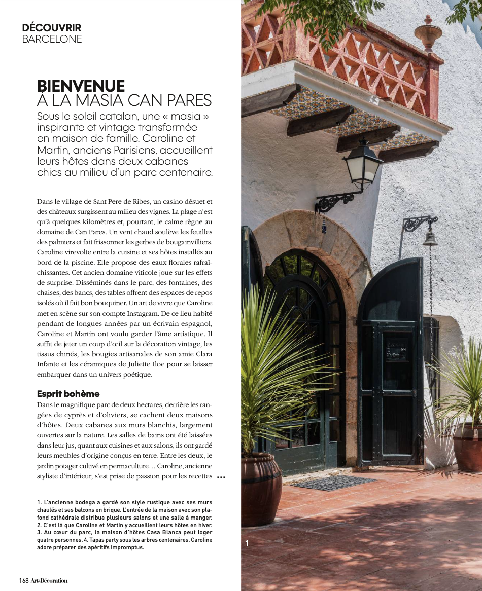 Portail d'une maison blanche avec un arceau en pierre, petits balcons en fer forgé, porte en bois noire, plantes en pot, et un balcon en terre cuite avec une balustrade en brique rouge. Fenêtres avec volets vert foncé.