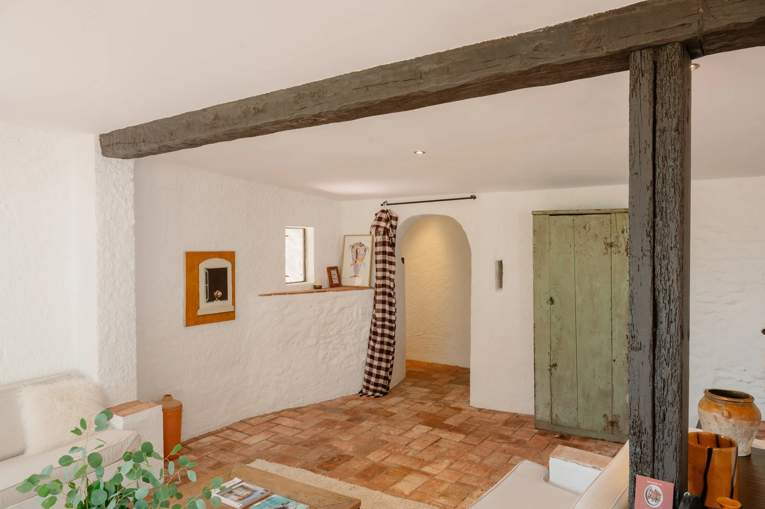 Interior of a rustic living room with white textured walls, a brick floor, a wooden beam ceiling, and simple furniture including a beige sofa, potted plants, and decorative vases.