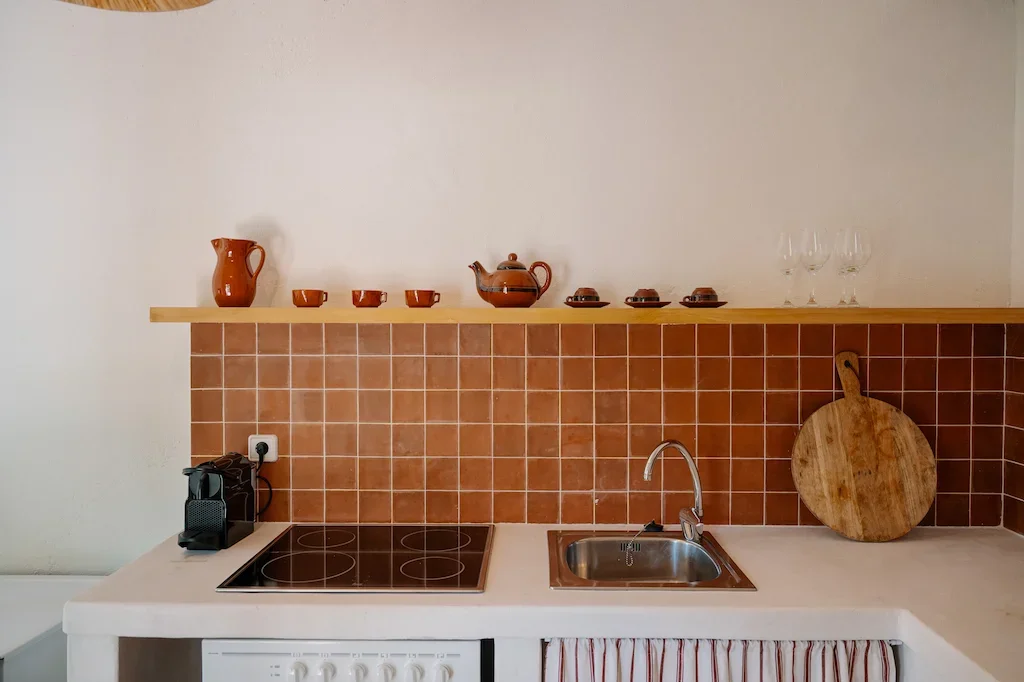 Cuisine avec une plaque de cuisson électrique, un évier, une robinetterie, une prise électrique, une cafetière, un plan de travail blanc, une crédence en carreaux rouges, une étagère en bois avec des vaisselles en céramique marron, un cercle en bois sur le plan de travail, et des verres en verre à vin sur l'étagère.