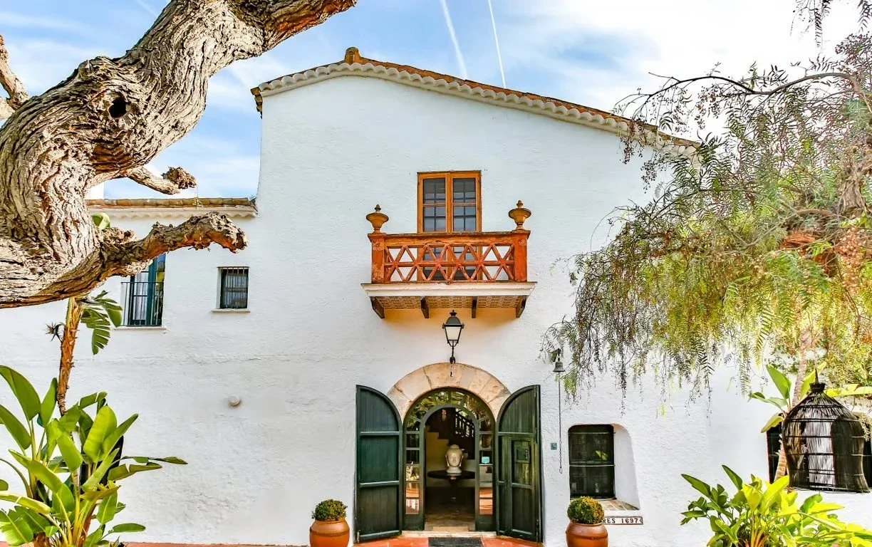 Maison blanche avec porte en arc, balcon en bois, entourée d'arbres et de plantes