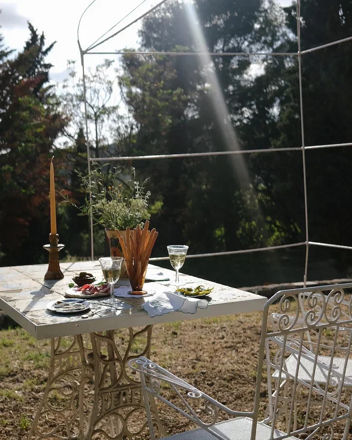 Table de jardin décorative avec une bougie, deux verres de vin, une plante en pot, des assiettes de nourriture, et une chaise en métal blanc dans un jardin en plein air.