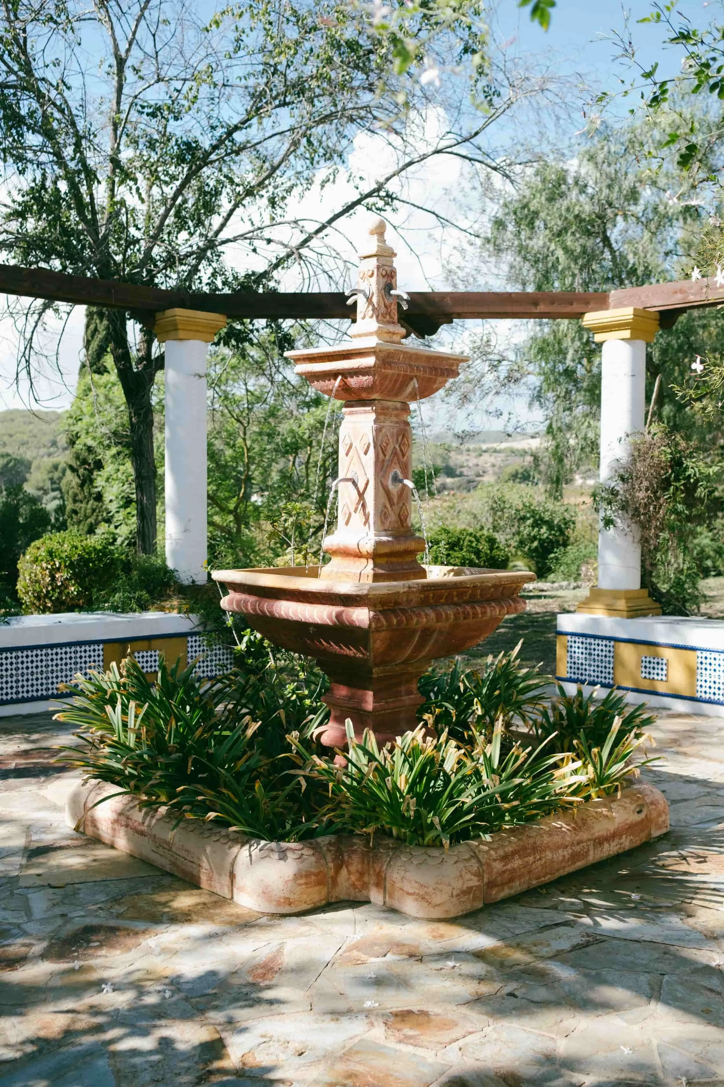 Pink and beige stone fountain with water spouts, surrounded by green plants, with a white and gold columned pergola in the background, under a partly cloudy sky.