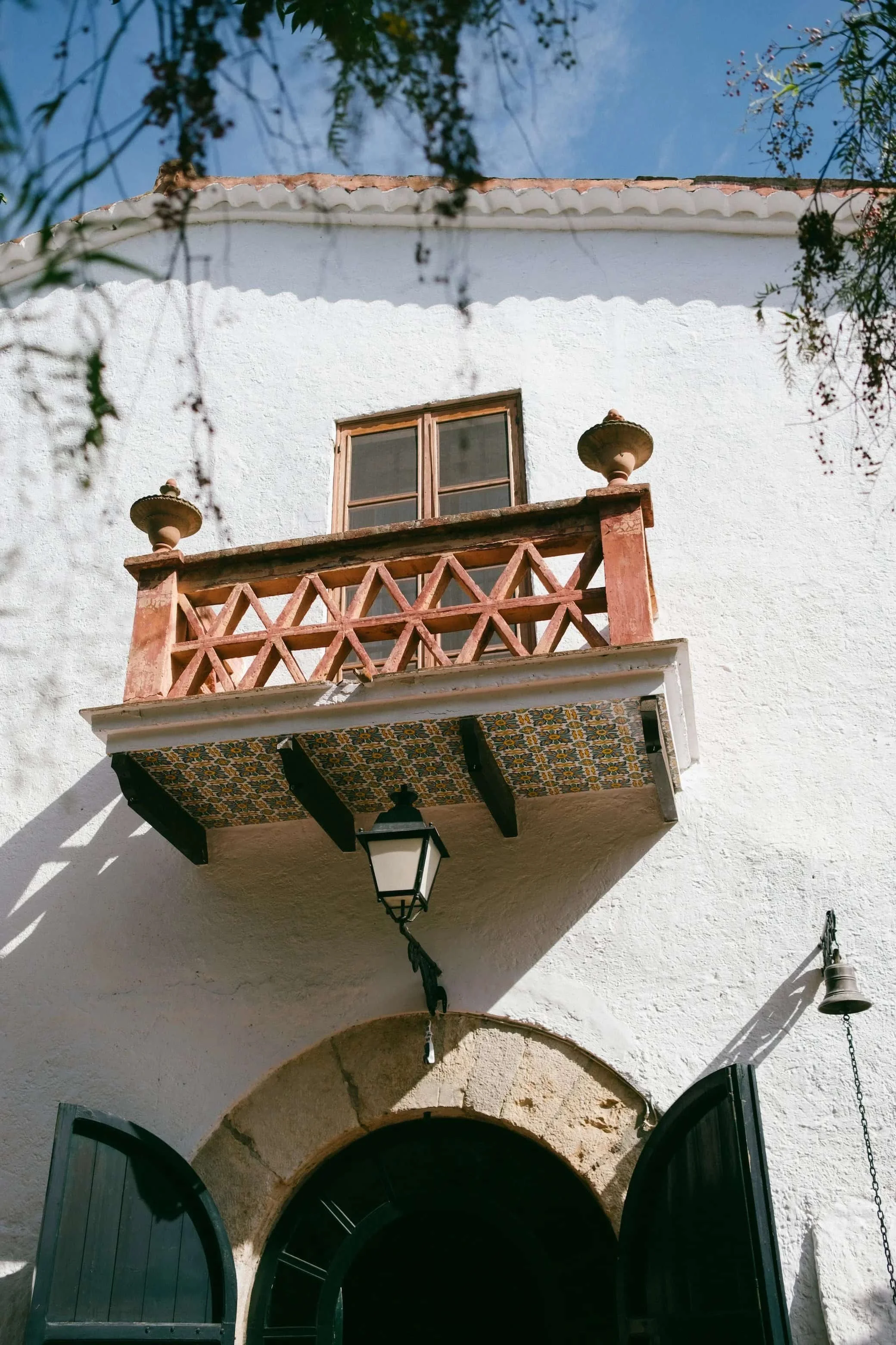 White stucco building with a small balcony, decorative tiled underside, black lantern, arched doorway, and wooden window, with tree branches and a blue sky above.