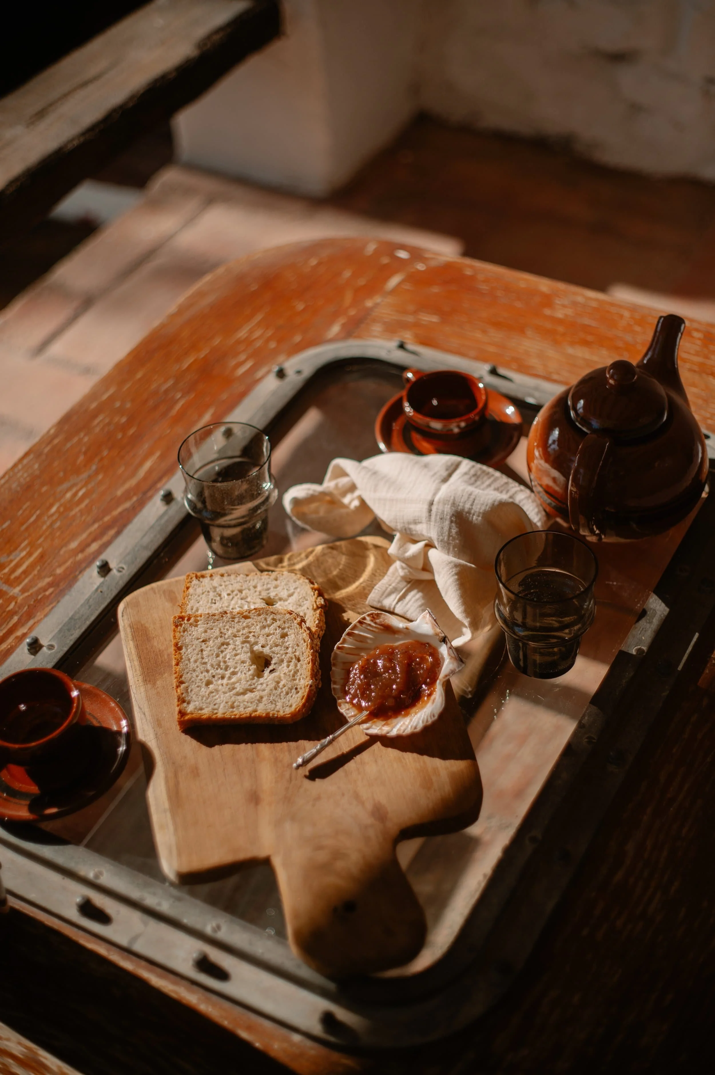 A wooden tray holding two slices of bread, a small spoon of jam, two glasses of water, and a brown teapot with two small cups, on a rustic wooden table.