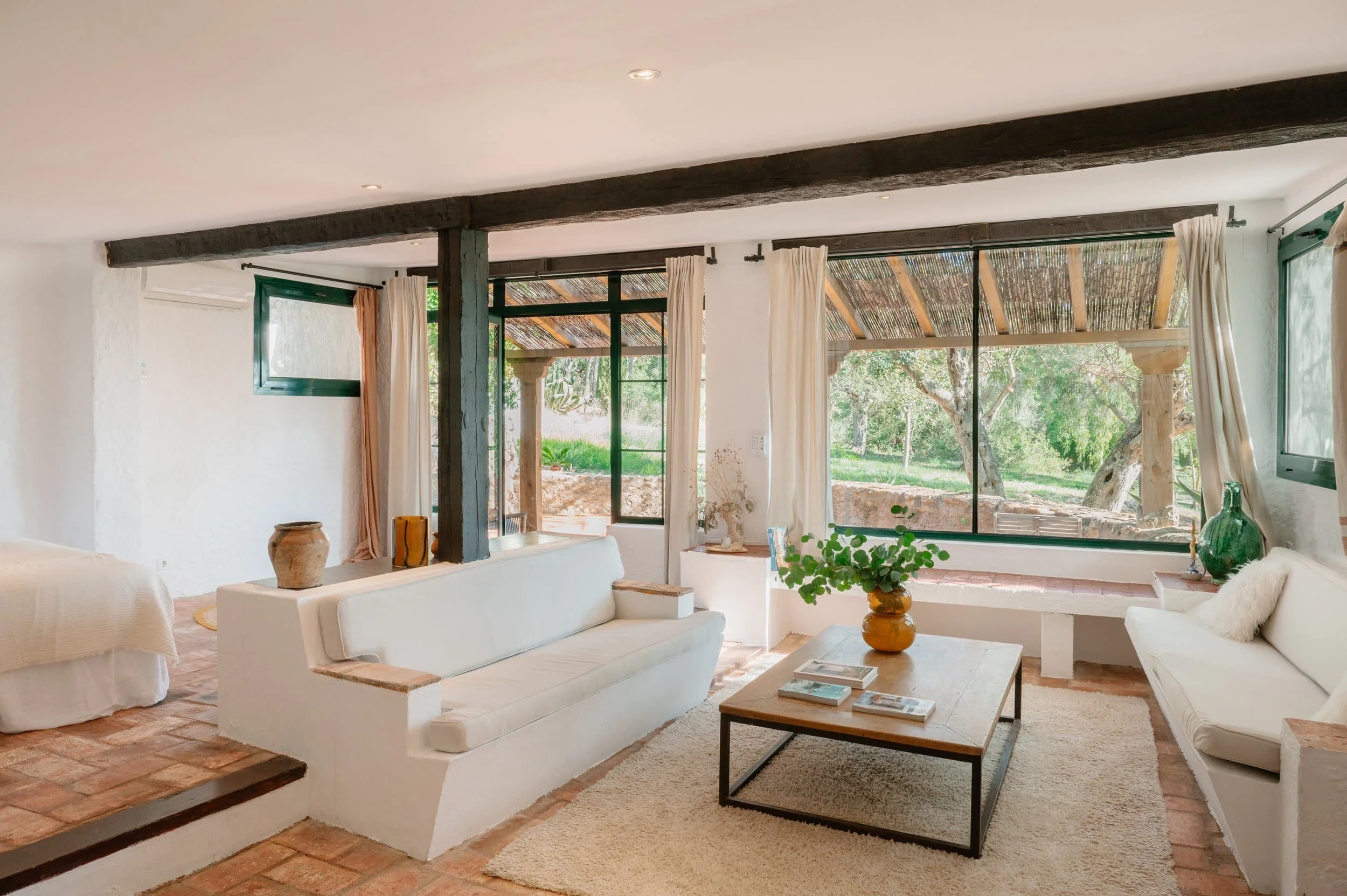 Living room with white couches, wooden coffee table, large windows, and greenery outside.