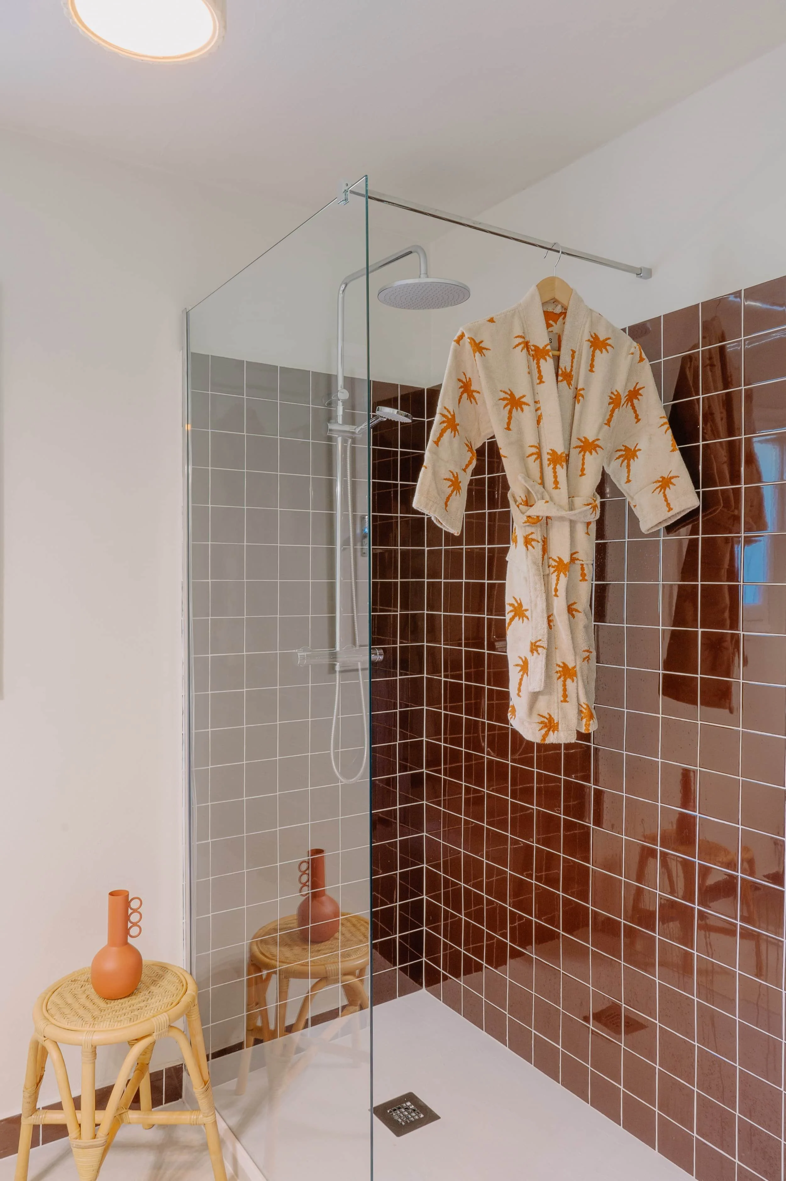 A bathroom shower area with brown tiled wall, a rain shower head, and a beige bathrobe with orange palm tree prints hanging on a hook. There are two small woven stools with matching vases.